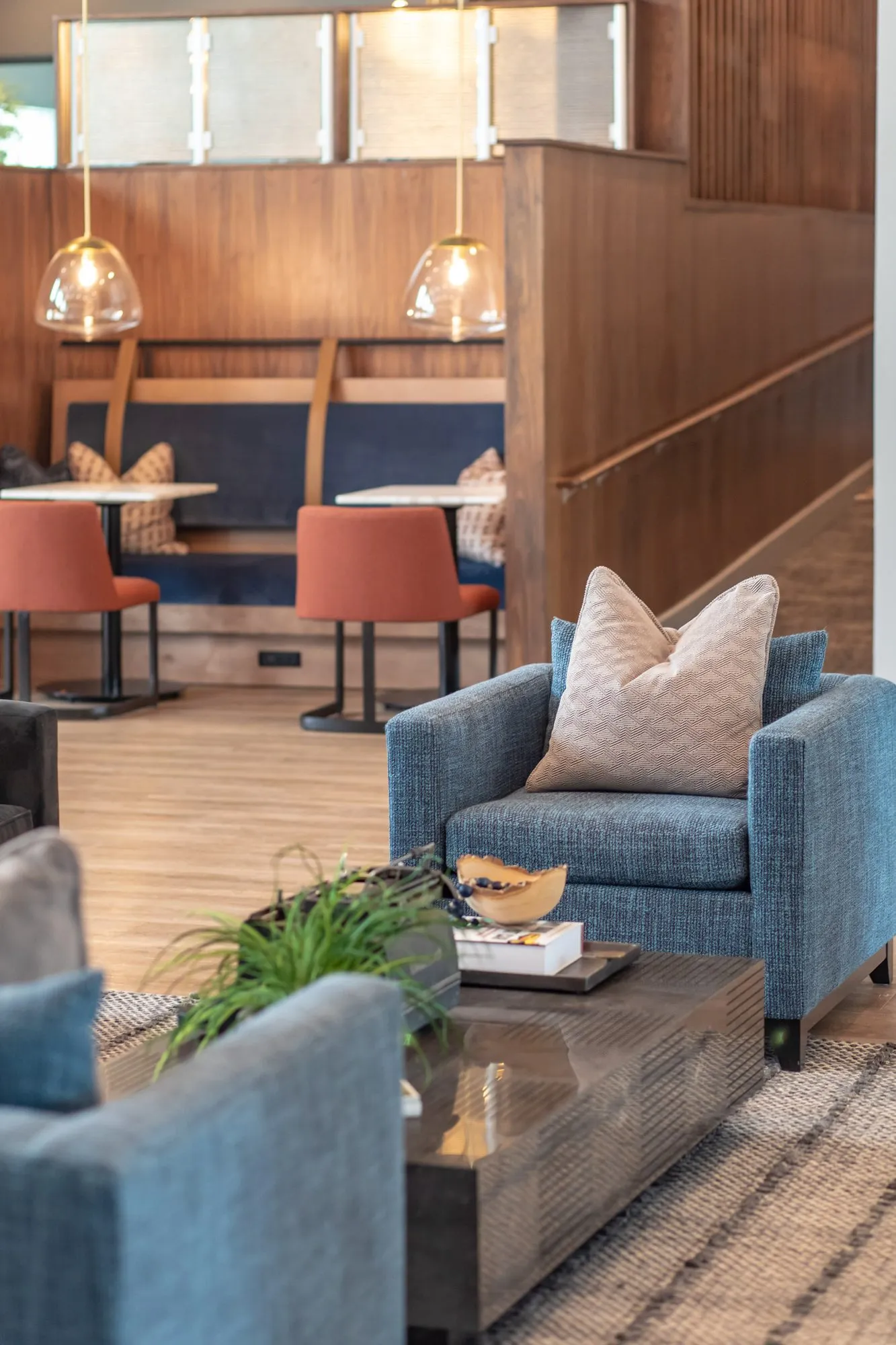 Multifamily amenity lounge with blue armchairs around a dark coffee table, walnut booths and rust chairs beyond