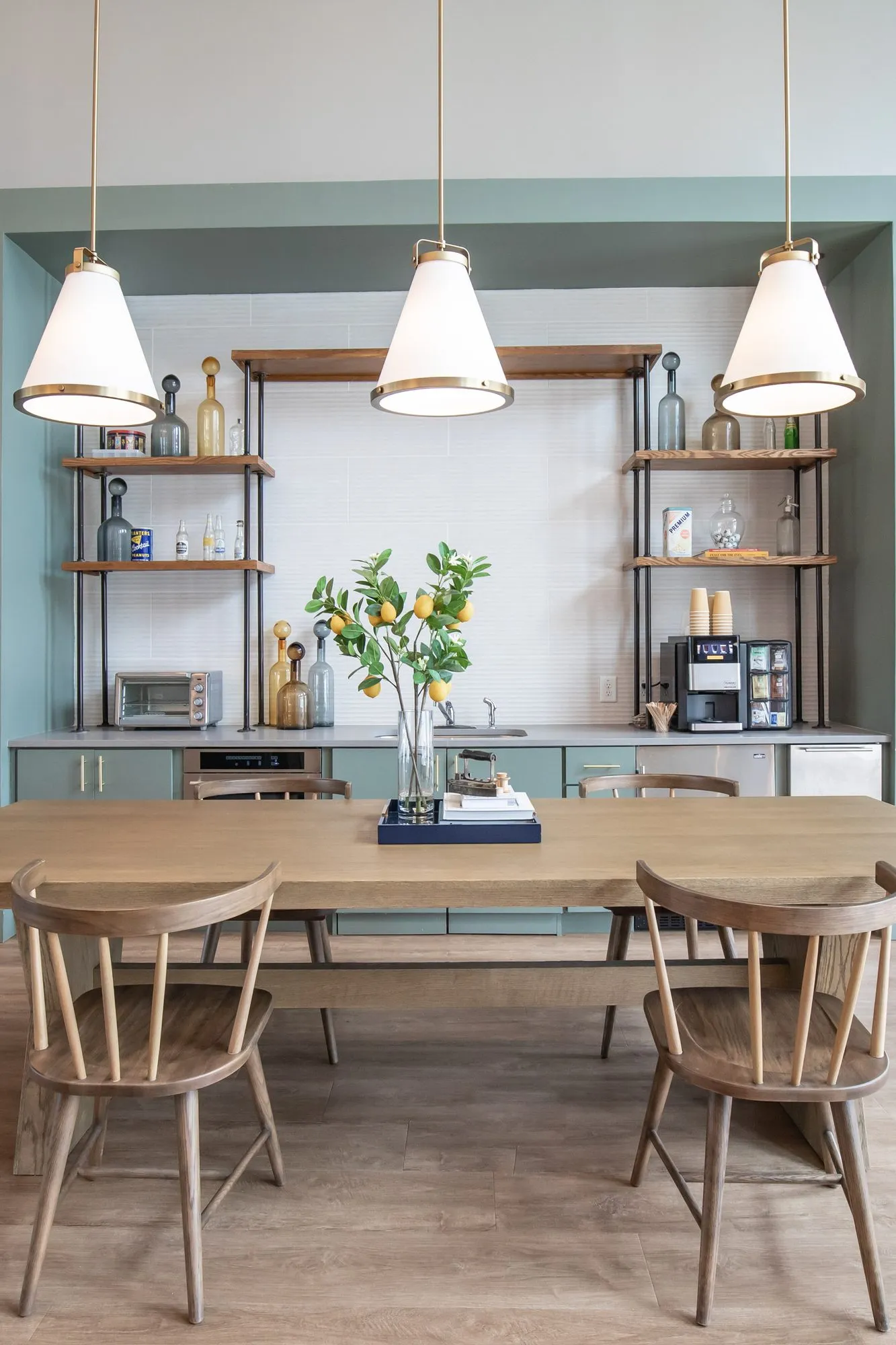 Multifamily amenity coffee bar with metal and wood shelving and cone pendants above a long table with spindle-back chairs