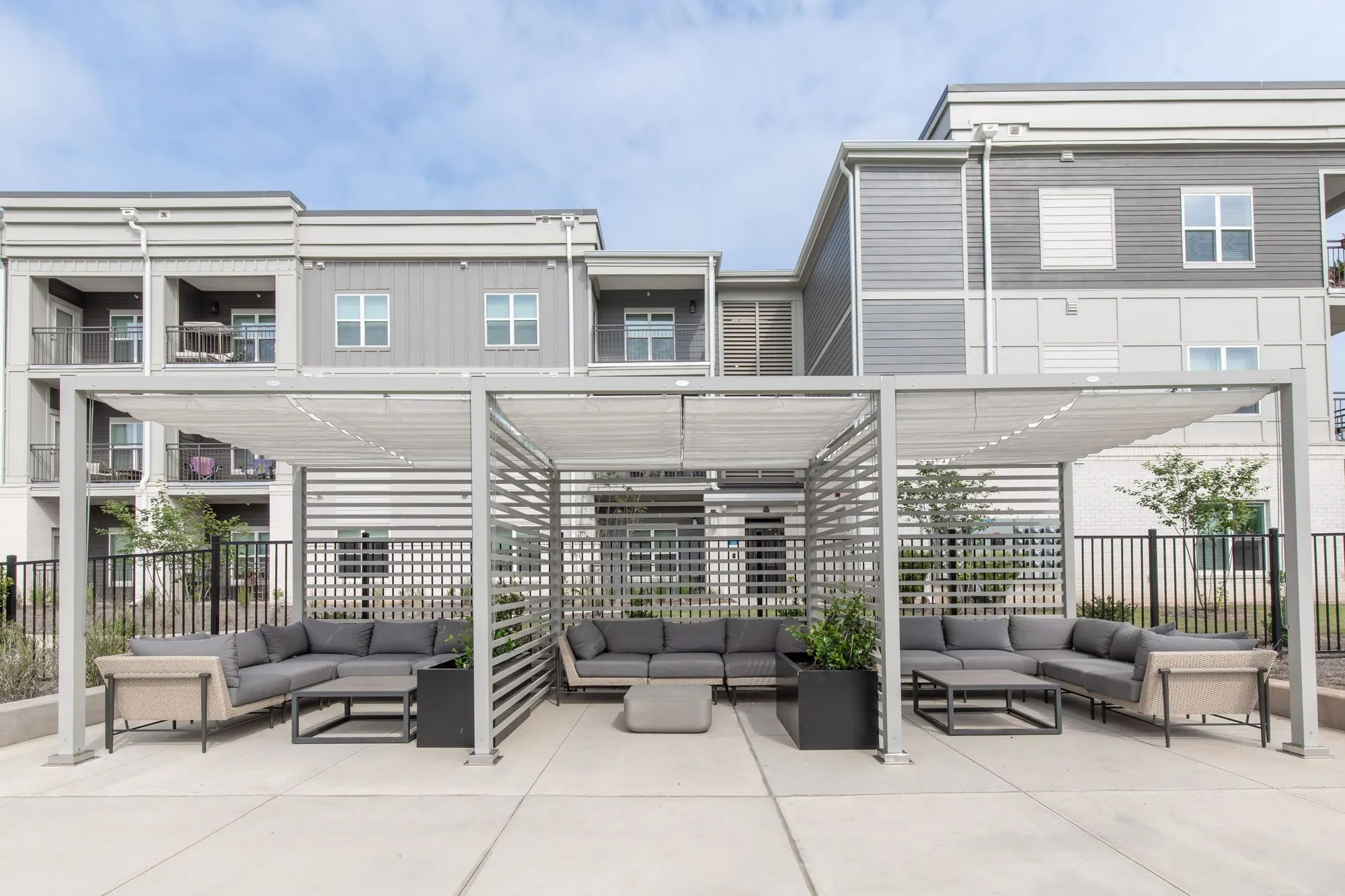 Multifamily community outdoor lounge under a white louvred pergola with grey sectional seating, apartment buildings behind