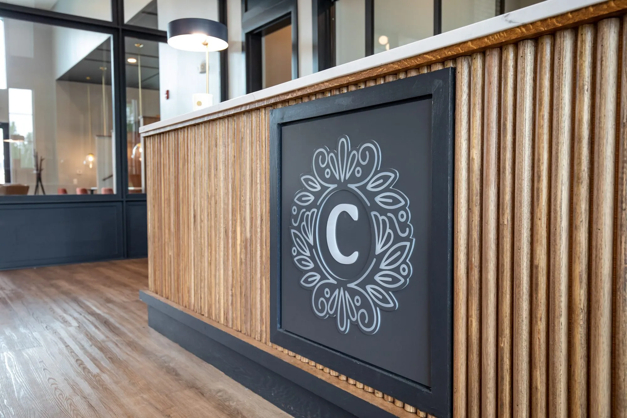 Multifamily amenity leasing desk clad in vertical wood dowels with a black panel bearing a white monogram C