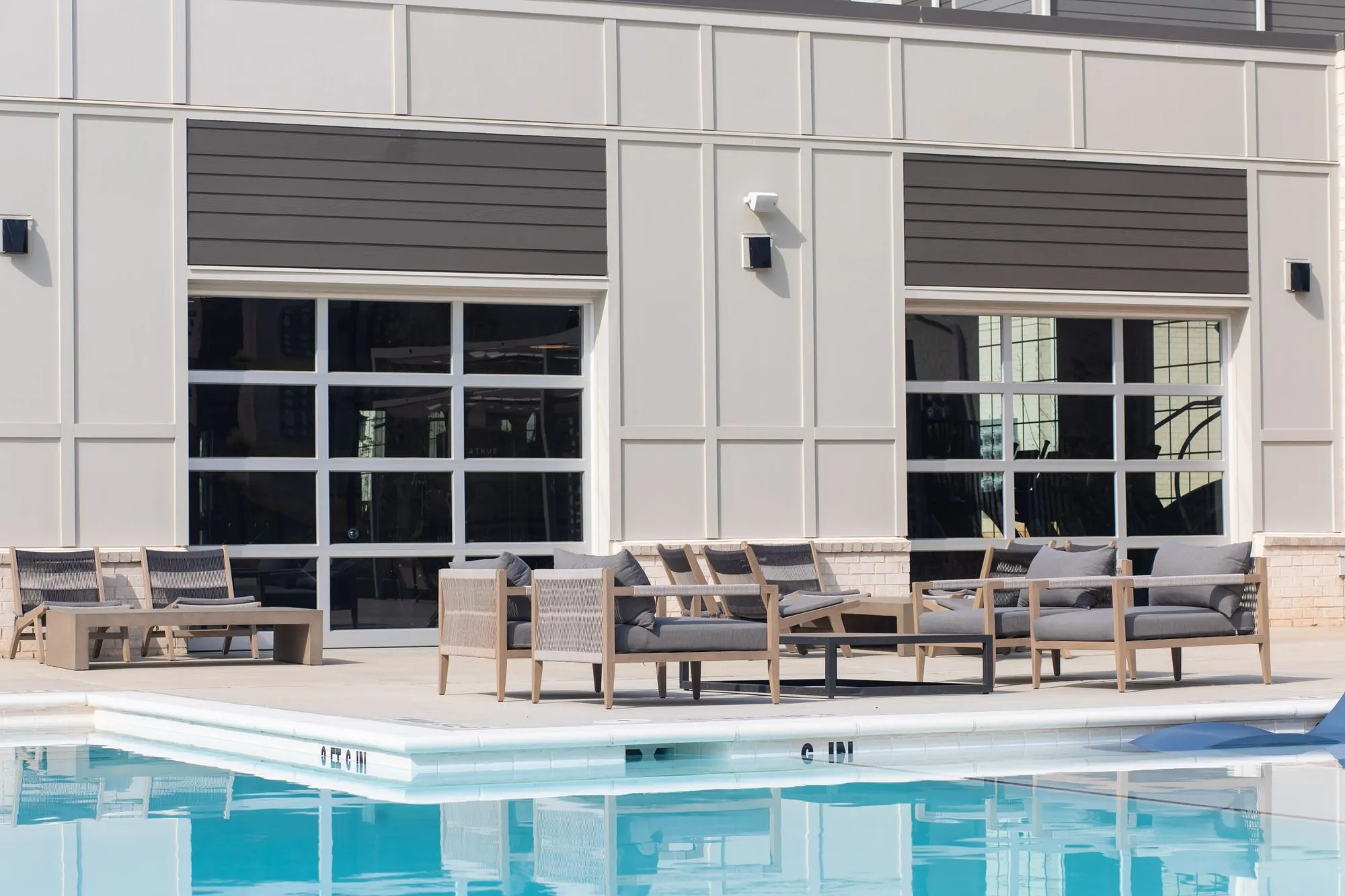 Multifamily community pool edge with teak and grey cushioned lounge chairs before the clubhouse's glass garage doors
