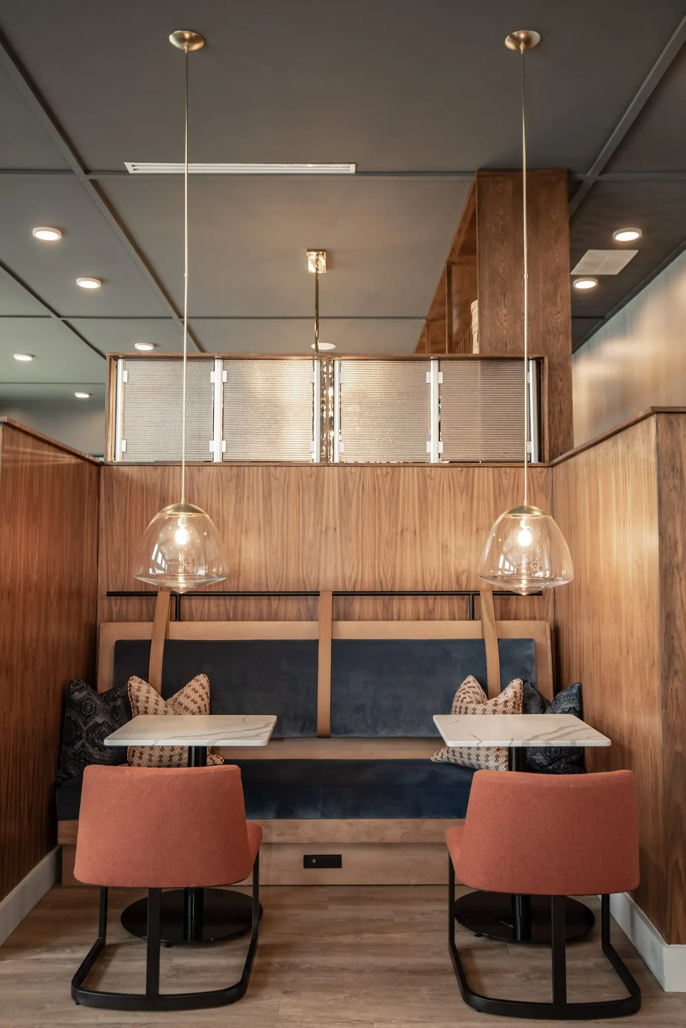 Multifamily amenity walnut booth with a navy bench and two rust chairs under a pair of glass pendants