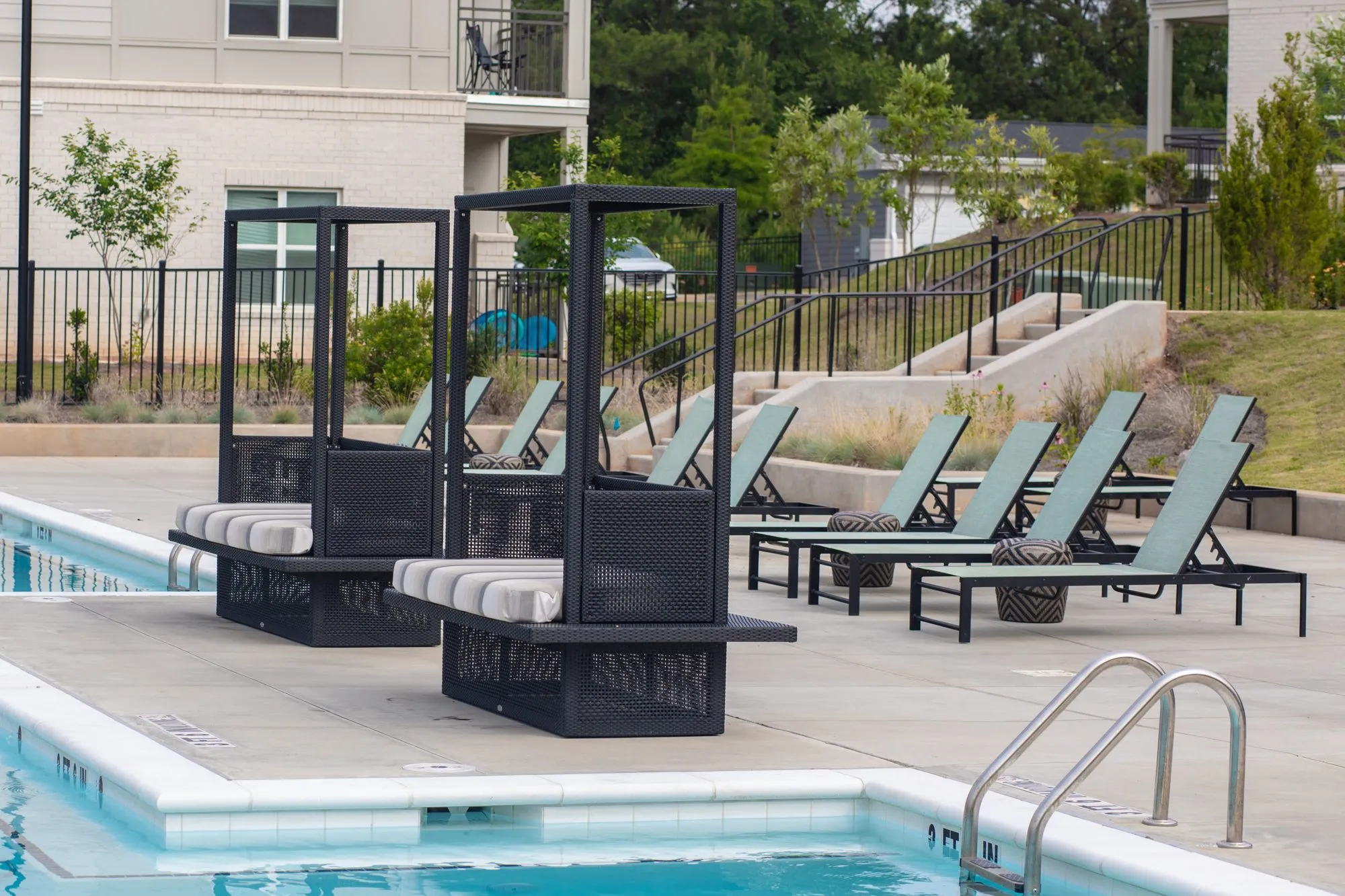 Multifamily community pool edge with black metal cabana frames and striped cushions, teal chaises on the deck behind