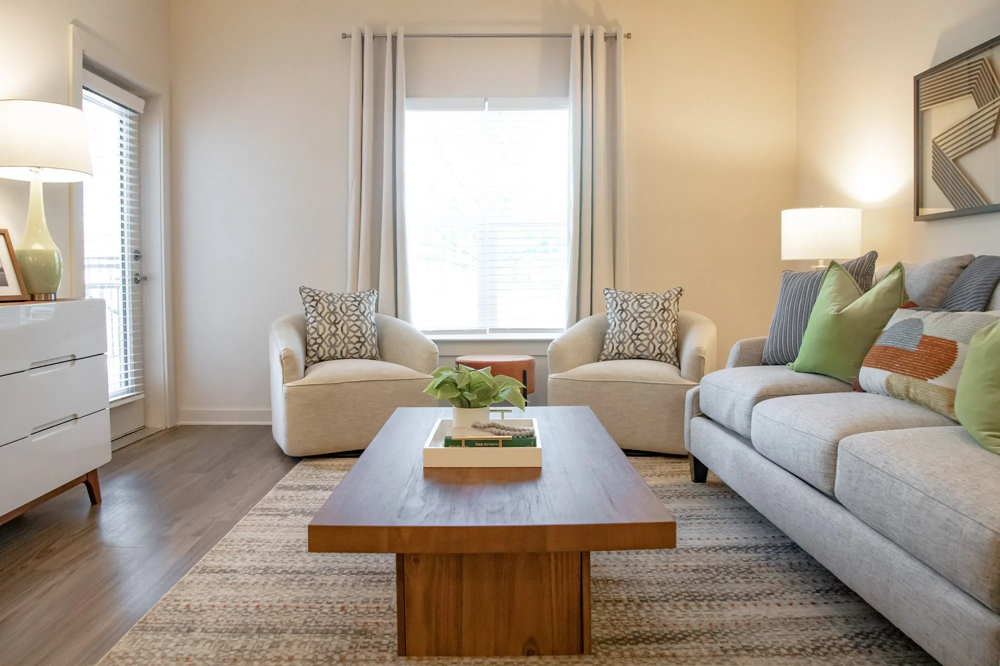 Multifamily apartment model living room with a grey sofa and green pillows, two cream armchairs and a block wood coffee table