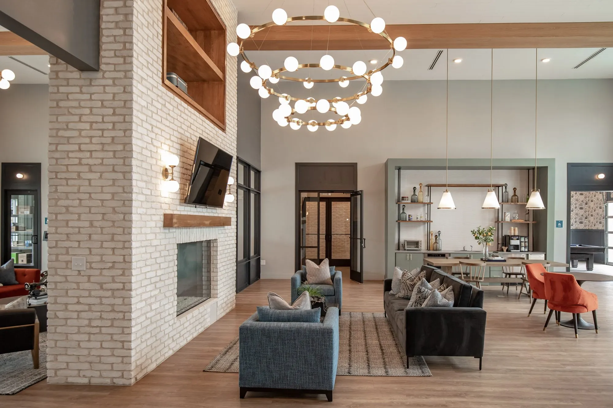 Multifamily amenity lounge with blue armchairs and a dark sofa beside the white brick fireplace, coffee bar beyond