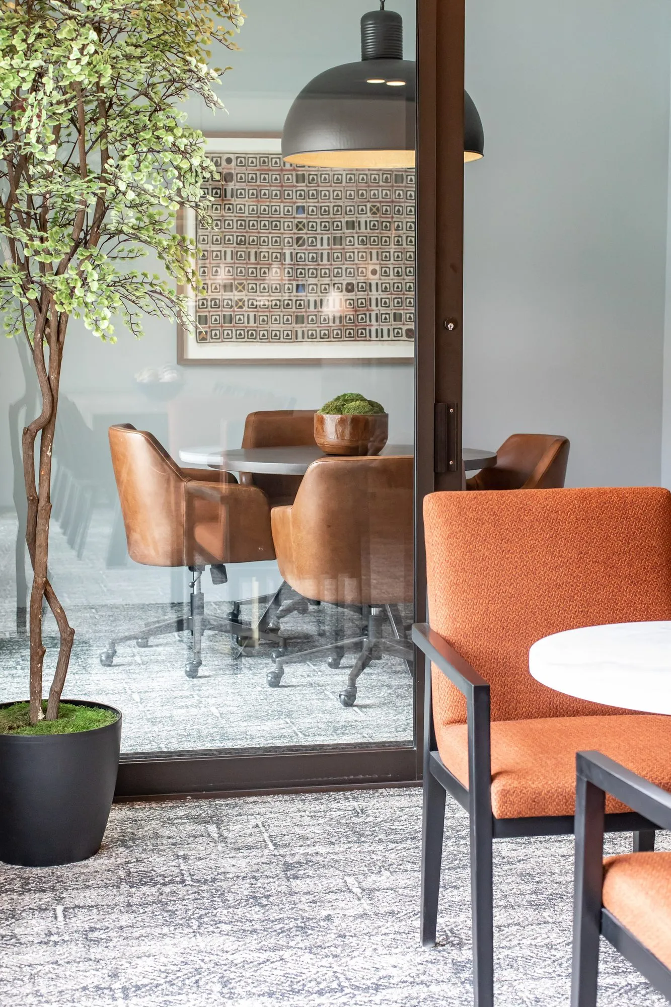 Multifamily amenity conference room seen through a glass wall, leather chairs at a round table, an orange chair in front