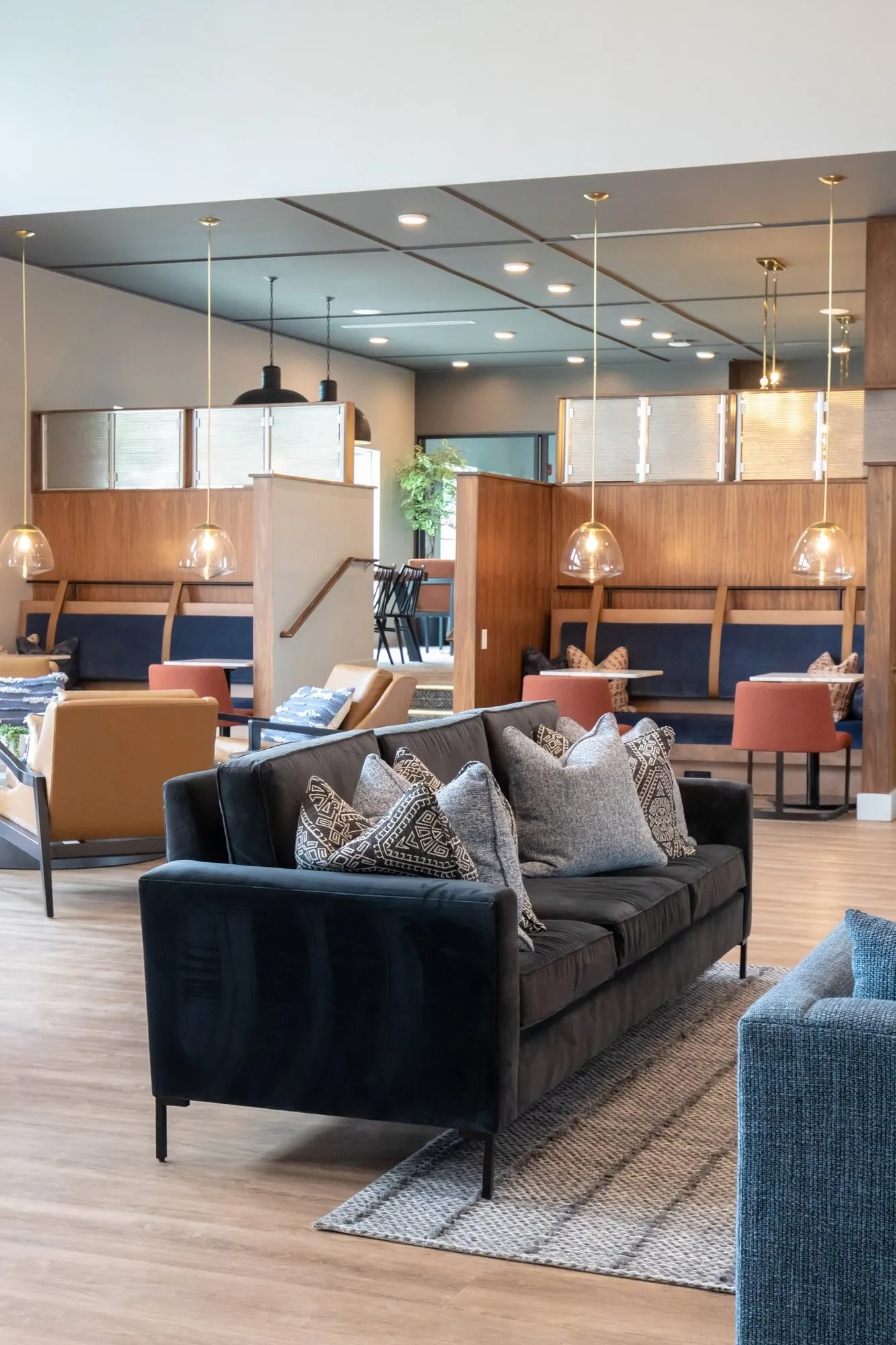 Multifamily amenity lounge seen past a dark velvet sofa toward walnut booths with rust chairs under glass pendants