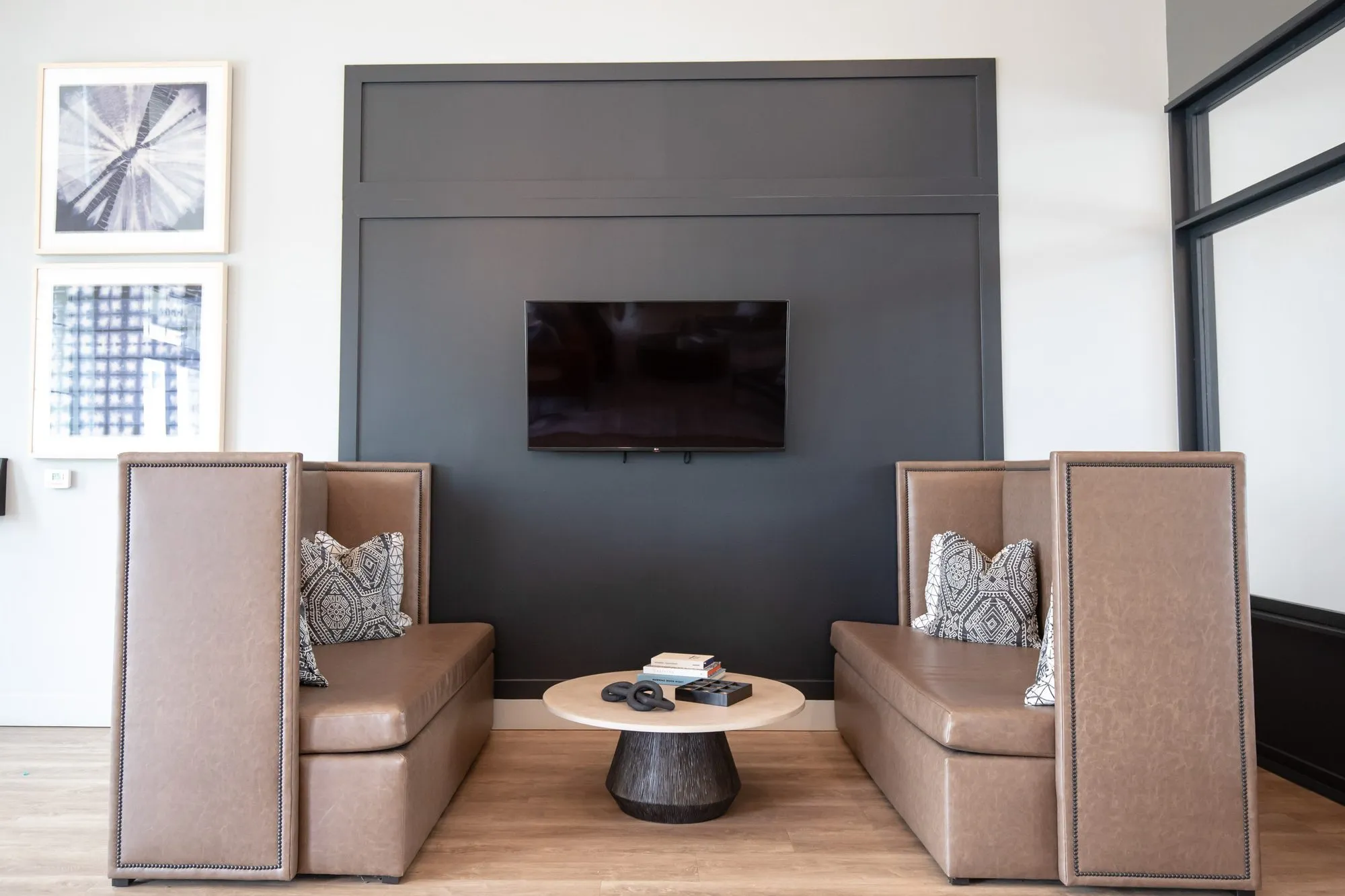 Multifamily amenity booth with two tan leather high-back seats across a round table before a dark panelled wall with a TV