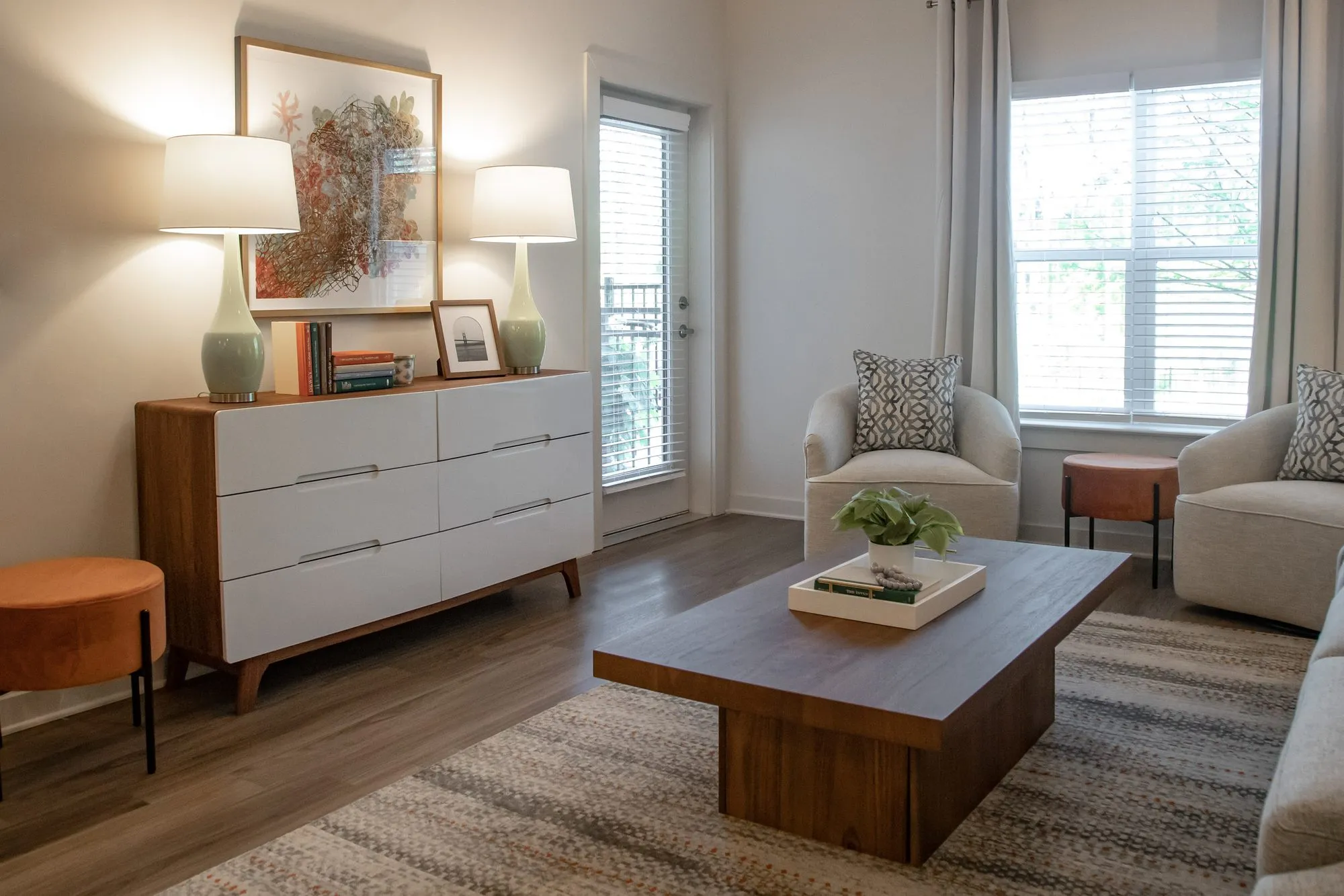 Multifamily apartment model living room with a white dresser between sage lamps, a dark coffee table and cream armchairs