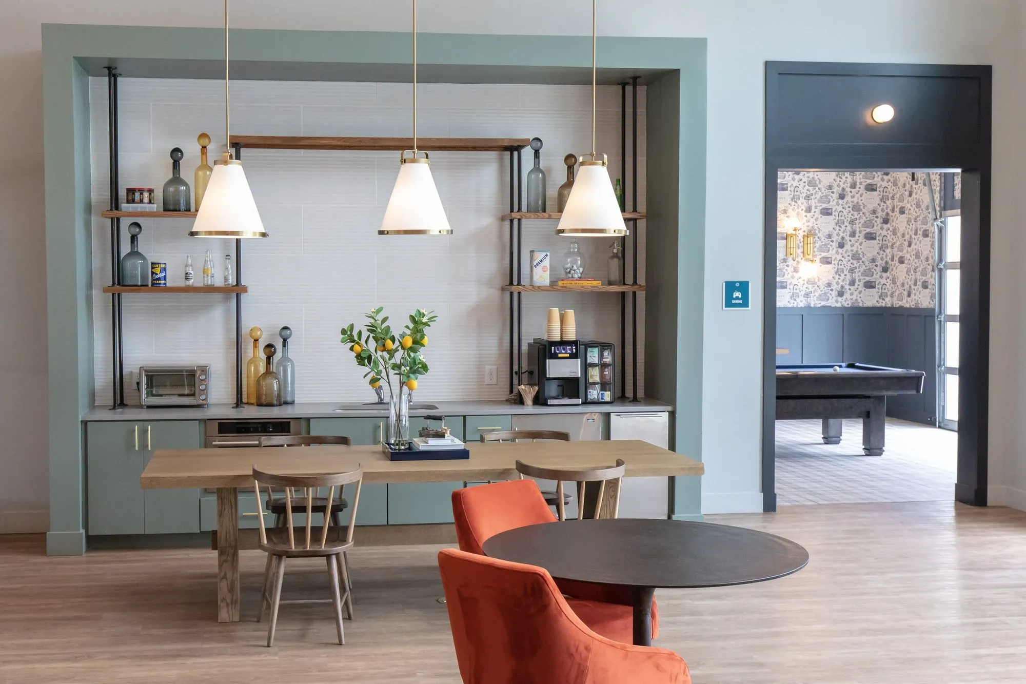 Multifamily amenity coffee bar with metal shelving and three cone pendants above a long wood table, a billiards room beyond