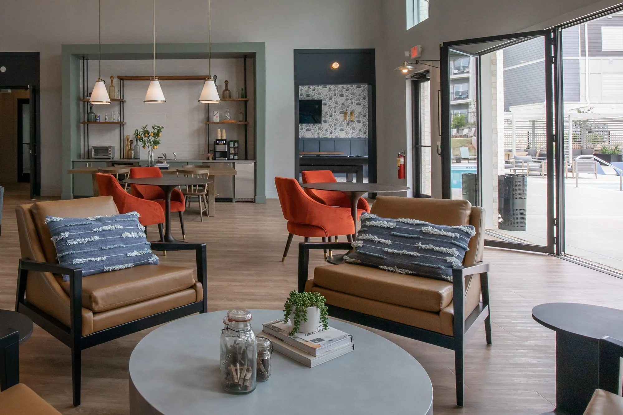 Multifamily amenity lounge with cognac leather armchairs around a grey round table, open glass doors to the pool deck
