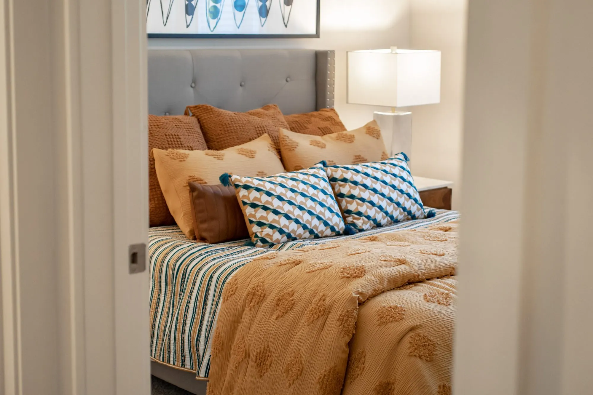 Multifamily apartment bedroom seen through a doorway, a grey tufted headboard with patterned pillows and a rust coverlet