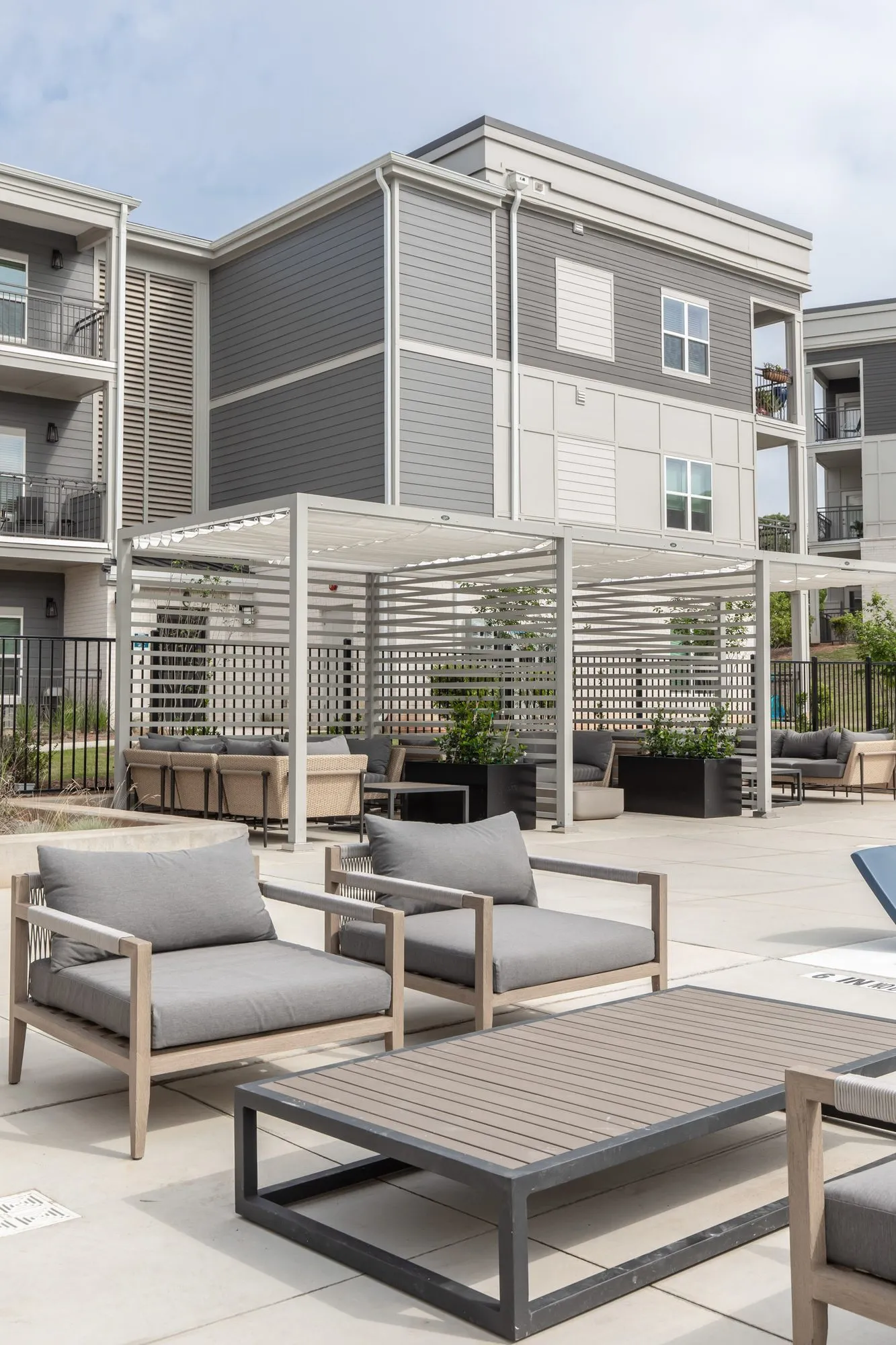 Multifamily community pool deck with grey-cushioned chairs around a wood coffee table, white louvred pergolas beyond