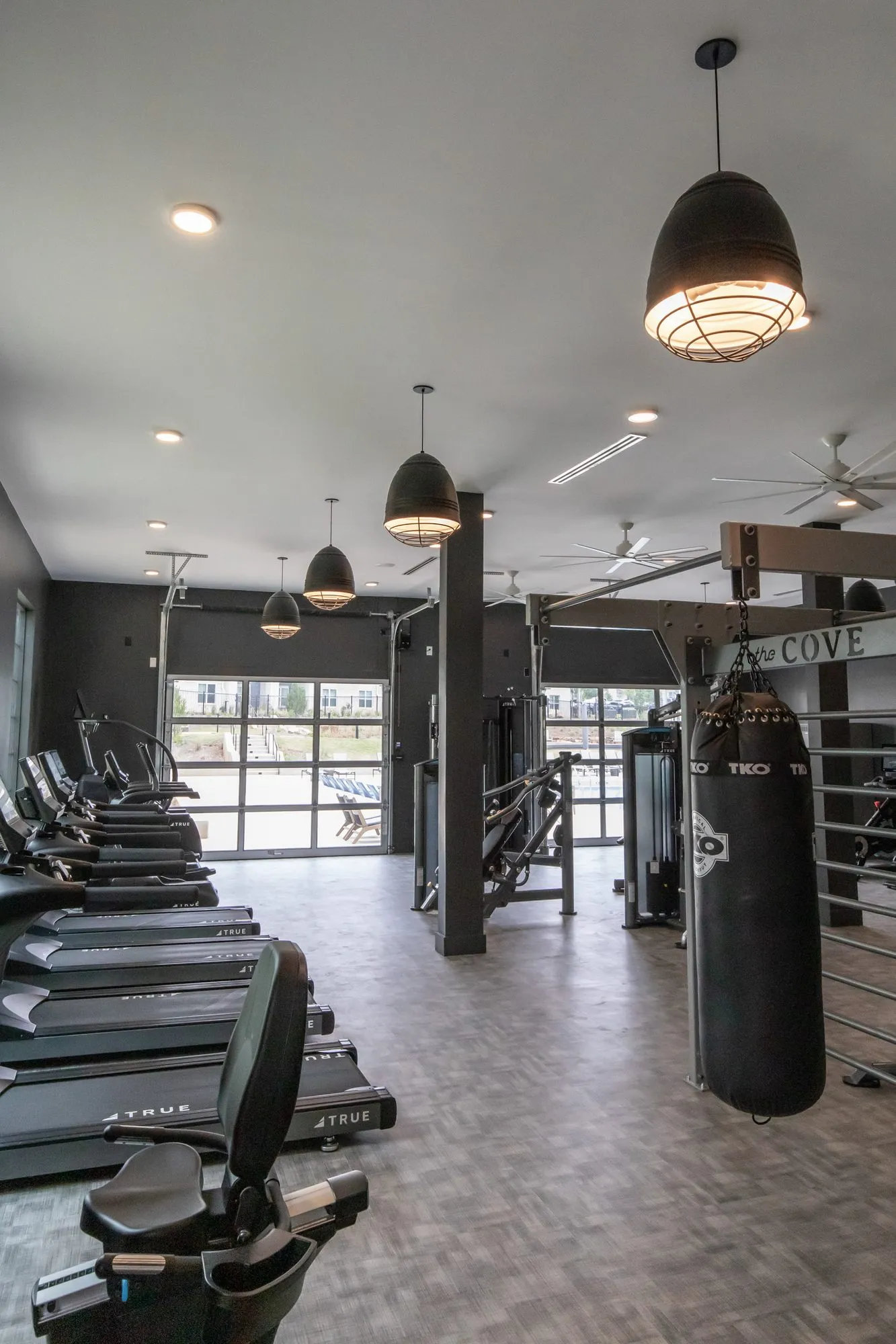 Multifamily amenity fitness center with treadmills and bikes along one side and a punching bag under black dome pendants