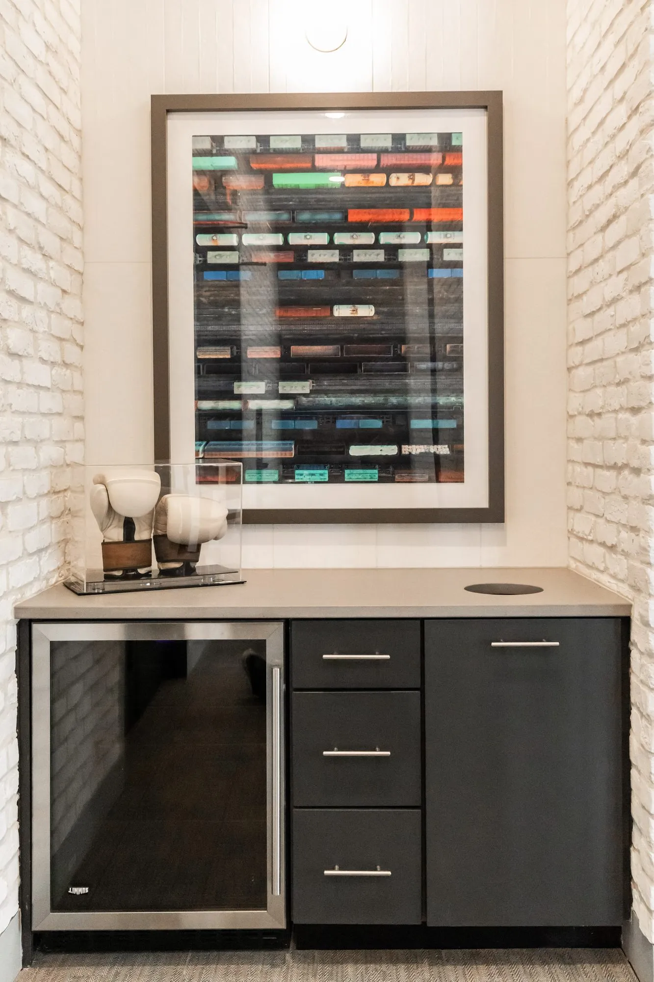 Multifamily amenity interior detail: dark cabinet with a beverage refrigerator below a framed abstract print of coloured bars