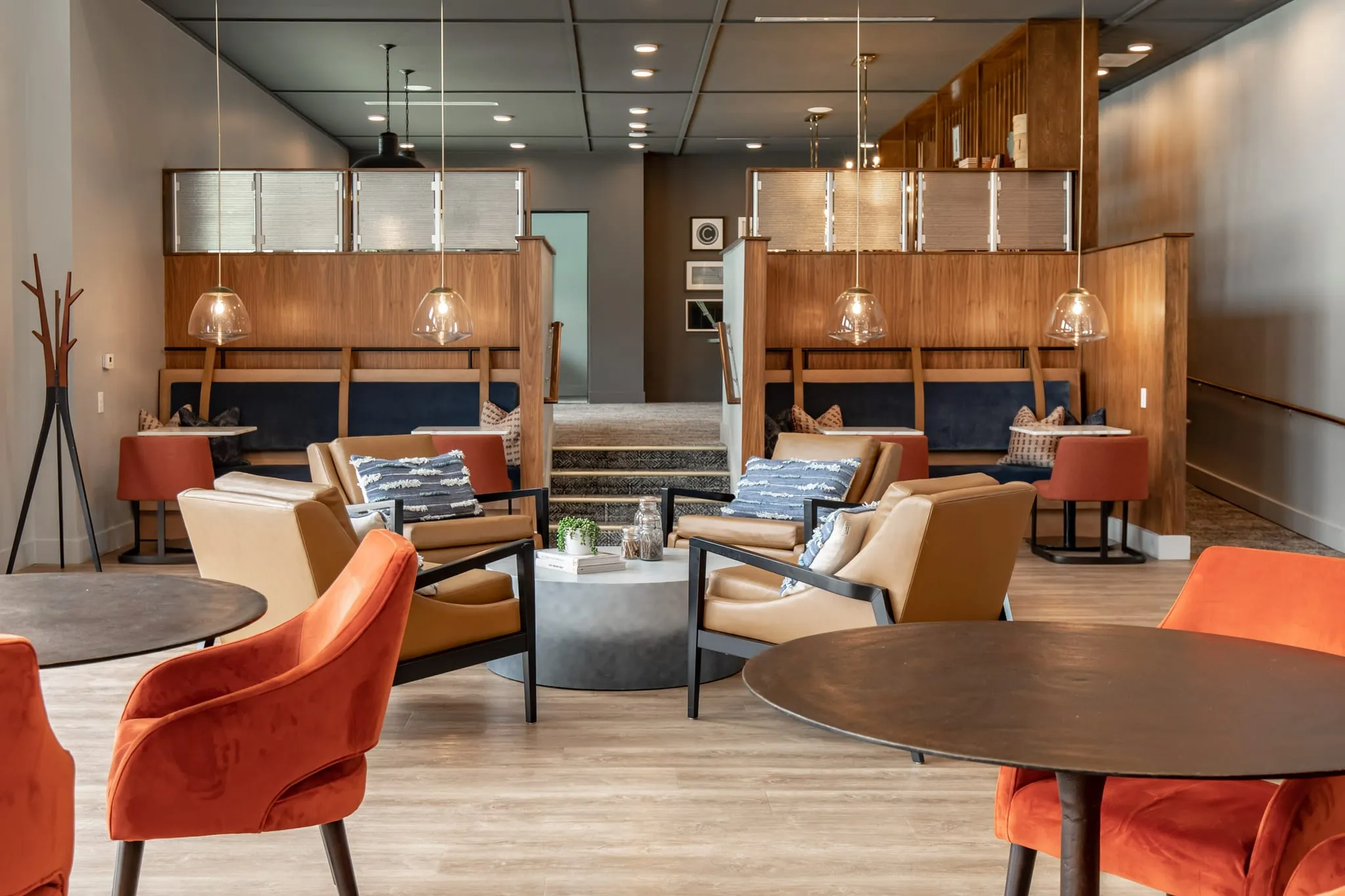 Multifamily amenity clubhouse lounge with orange armchairs around a round table, pendant-lit banquettes in millwork beyond