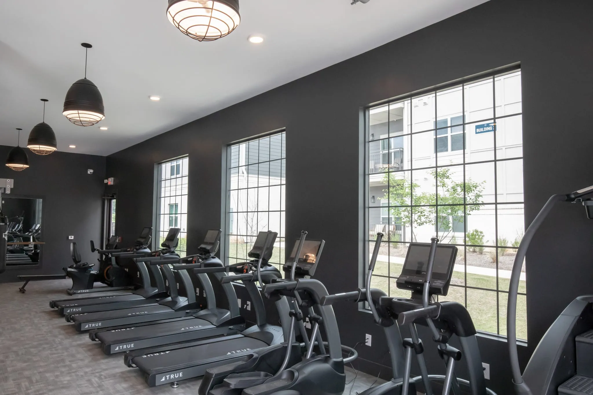 Multifamily amenity fitness center with treadmills along a charcoal wall under drum pendants beside tall windows