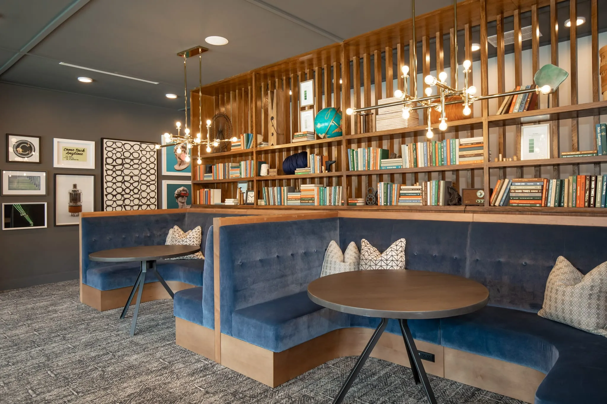 Multifamily amenity library banquette with navy velvet seating below a wall of bookshelves and wood slats lit by bare bulbs