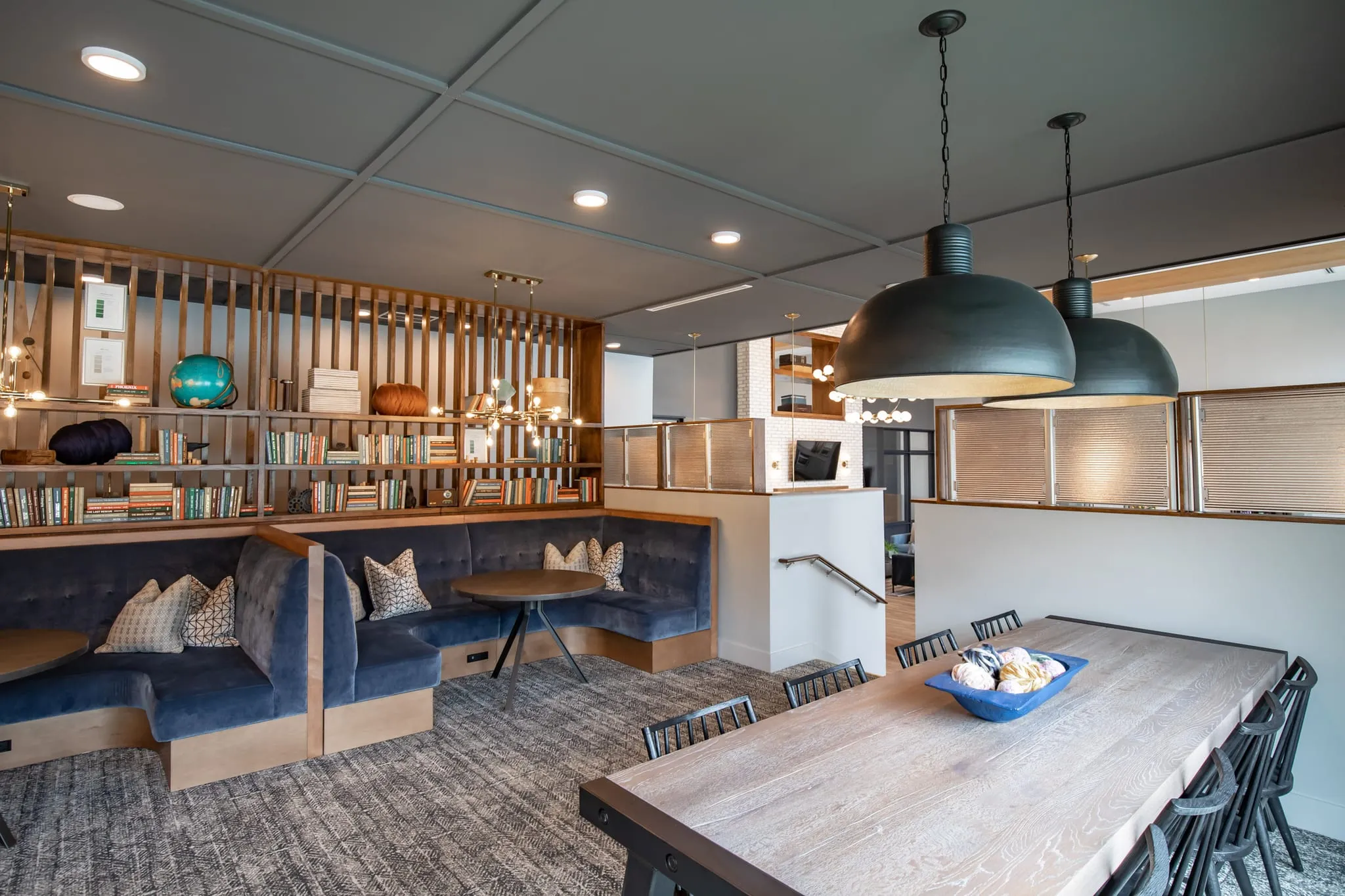 Multifamily amenity communal work area with navy banquettes and a long wood table under black dome pendants by bookshelves