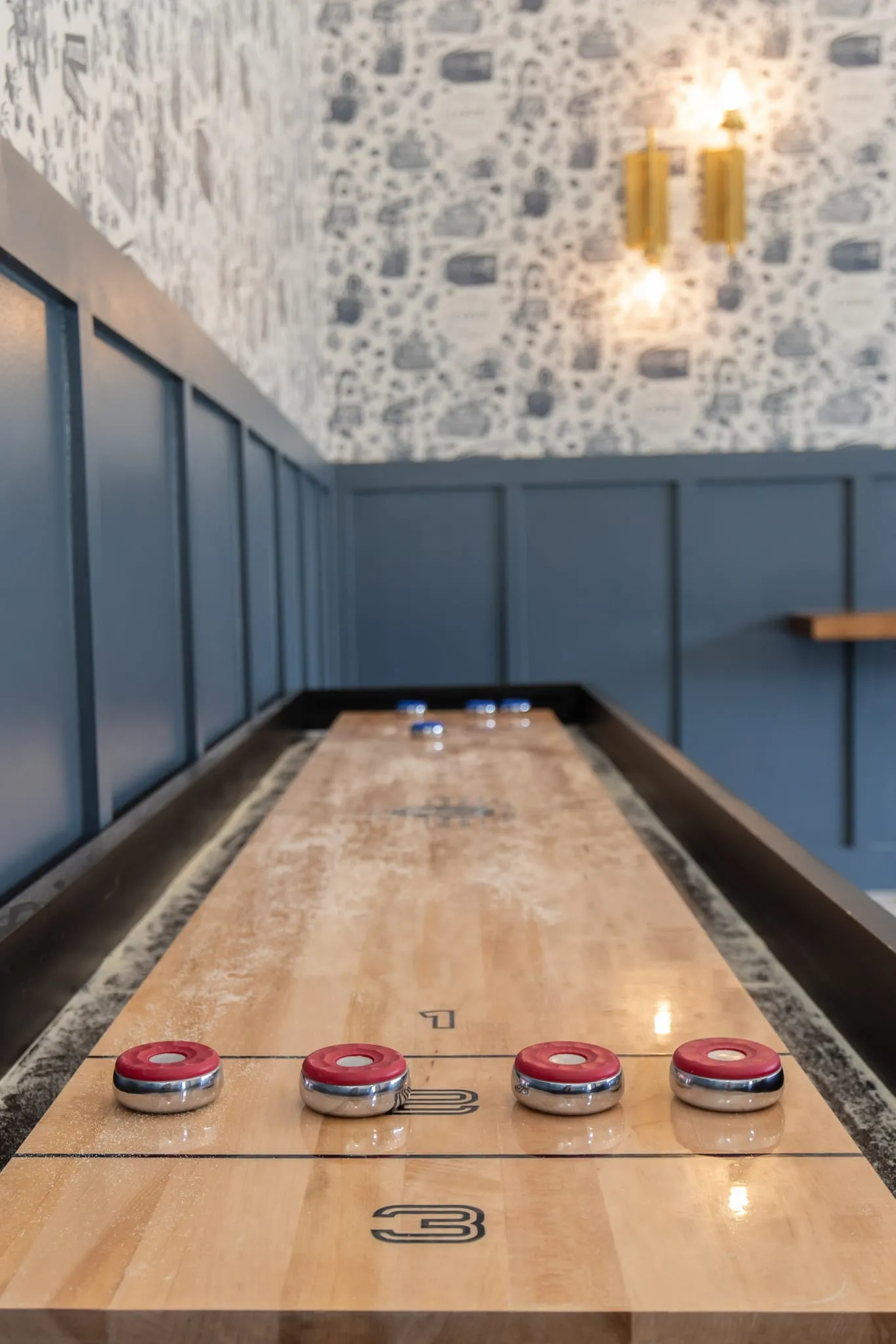 Multifamily amenity game room shuffleboard, pucks on the lane below brass sconces and toile wallcovering