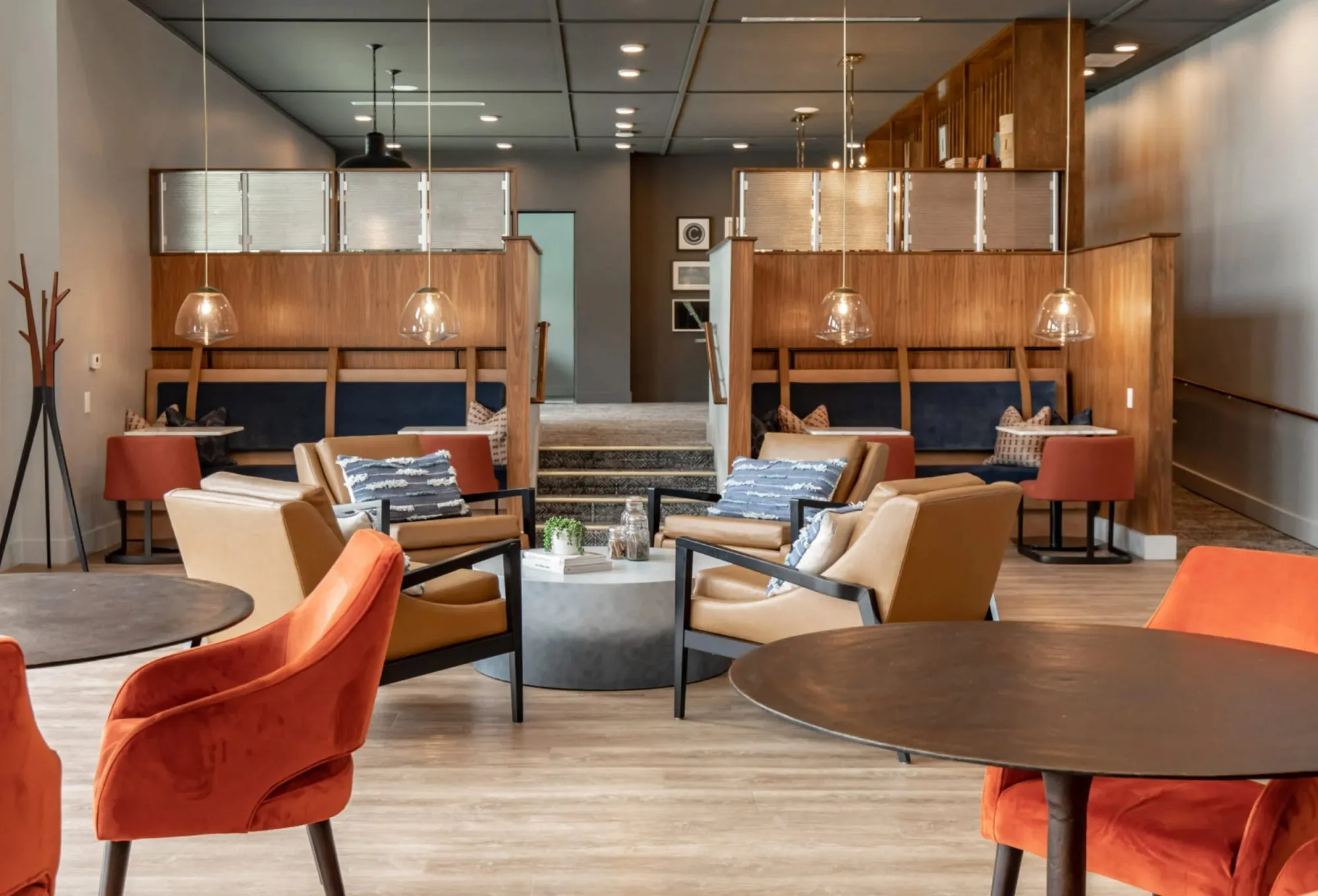 Coworking lounge with walnut booth banquettes, glass globe pendants, orange and tan armchairs and round bistro tables