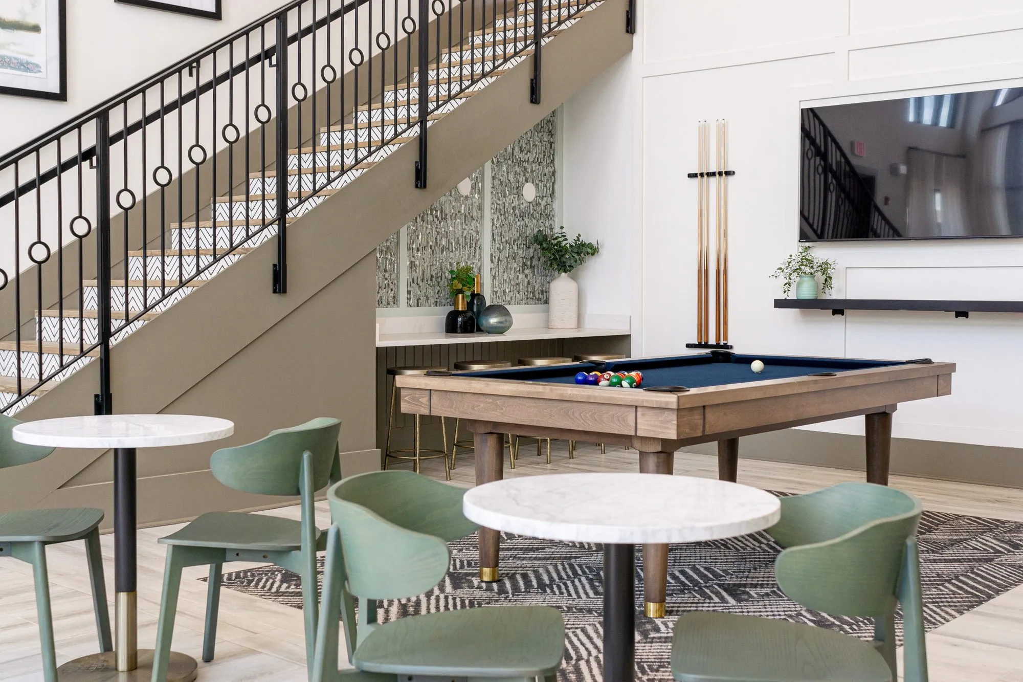 Multifamily amenity billiards table and sage cafe chairs beside the iron stair with circle balusters, a cue rack on the wall