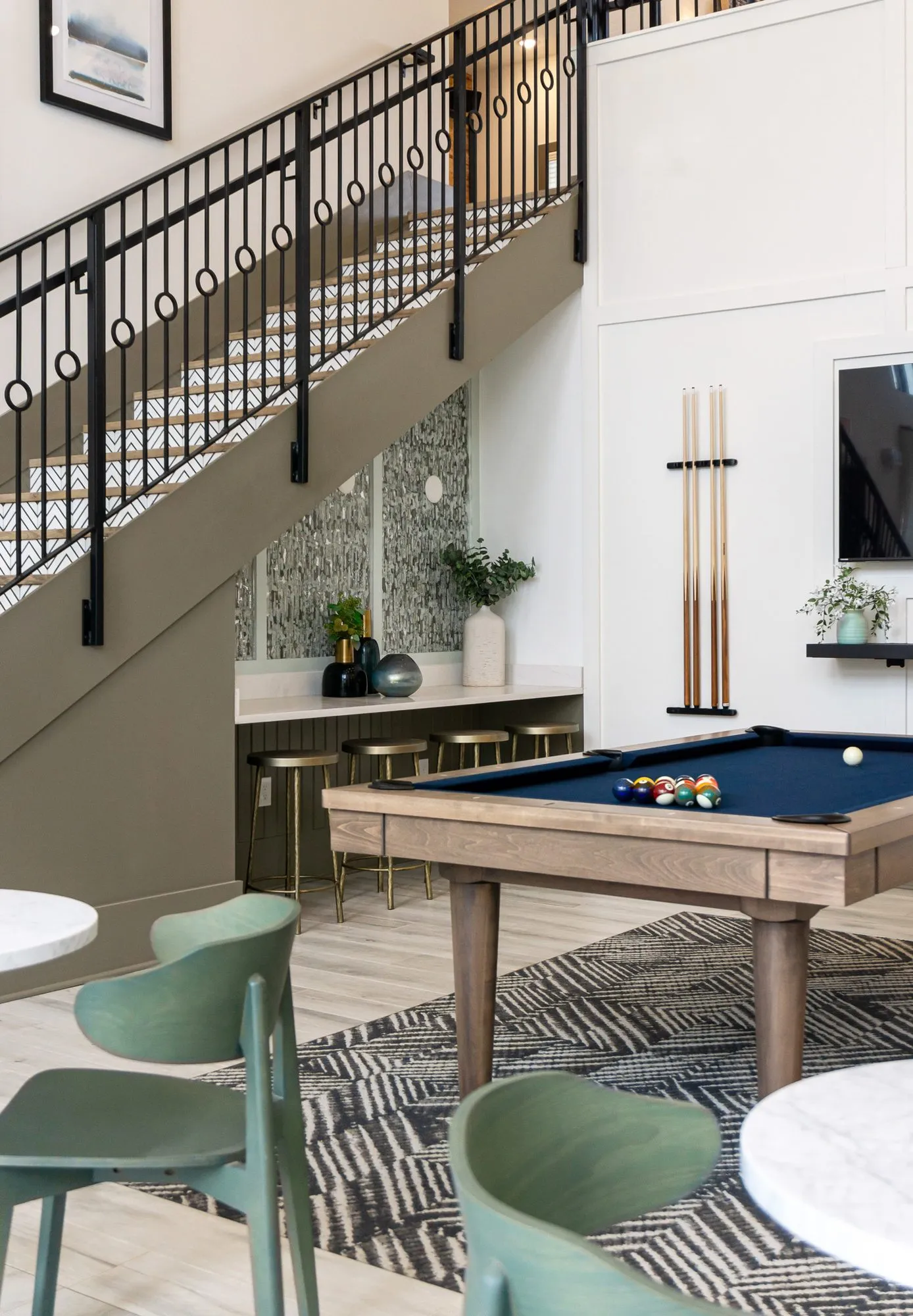 Multifamily amenity billiards table with racked balls before the stair alcove counter and brass stools, cues on the wall