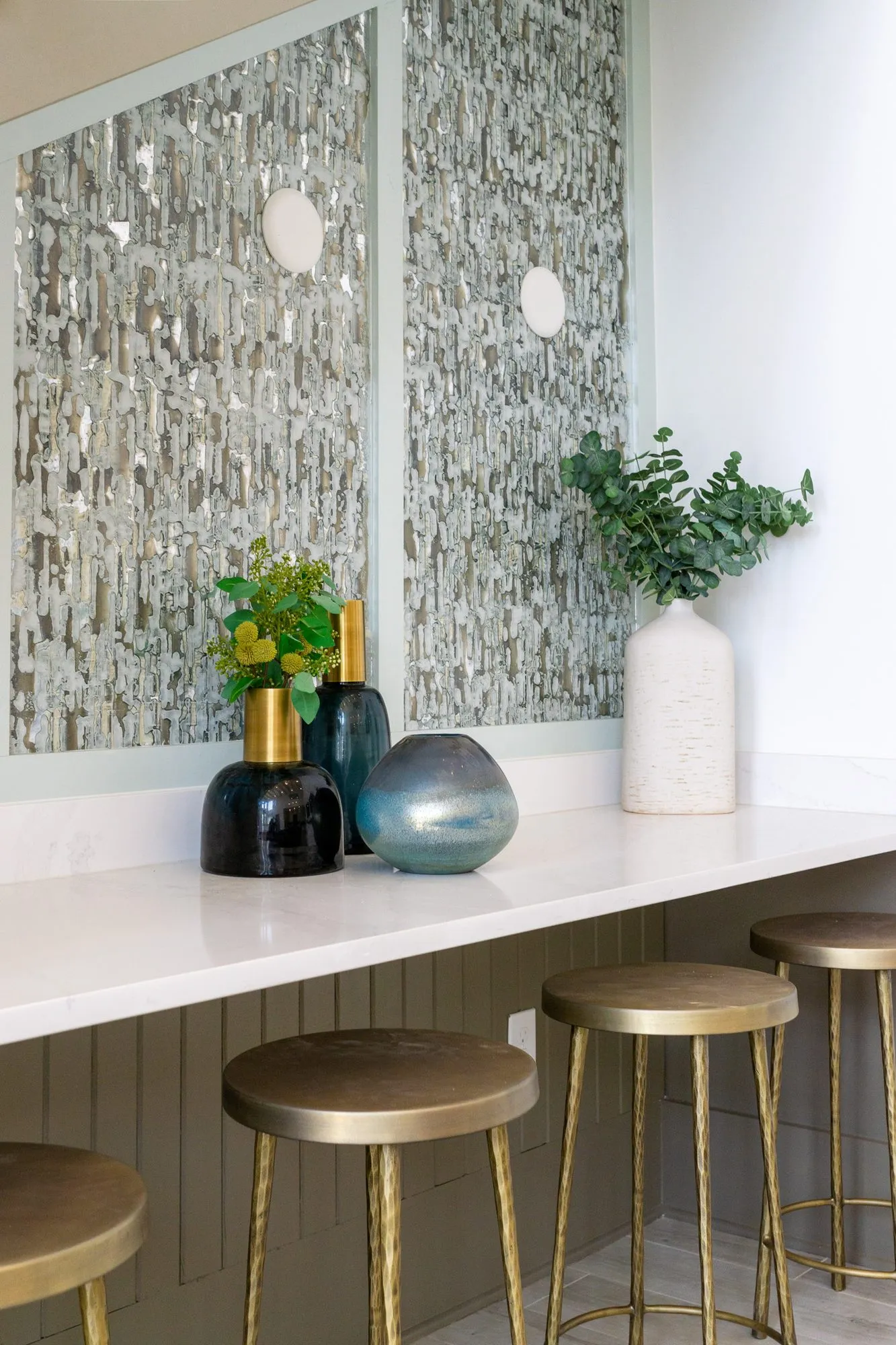 Multifamily interior detail: brass stools at a white counter below panels of textured metallic tile, with glass vases
