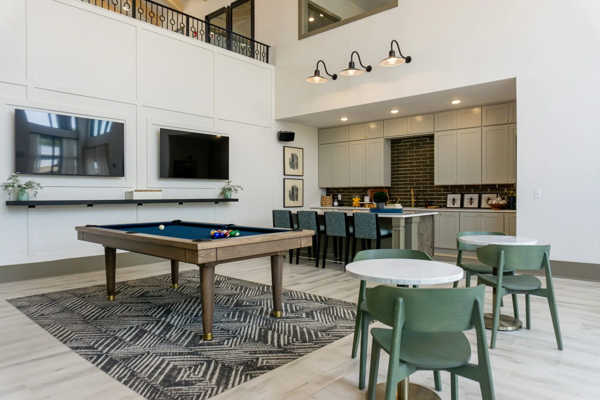 Multifamily amenity billiards table on a patterned rug before the double-height wall, kitchen with brick tile beyond