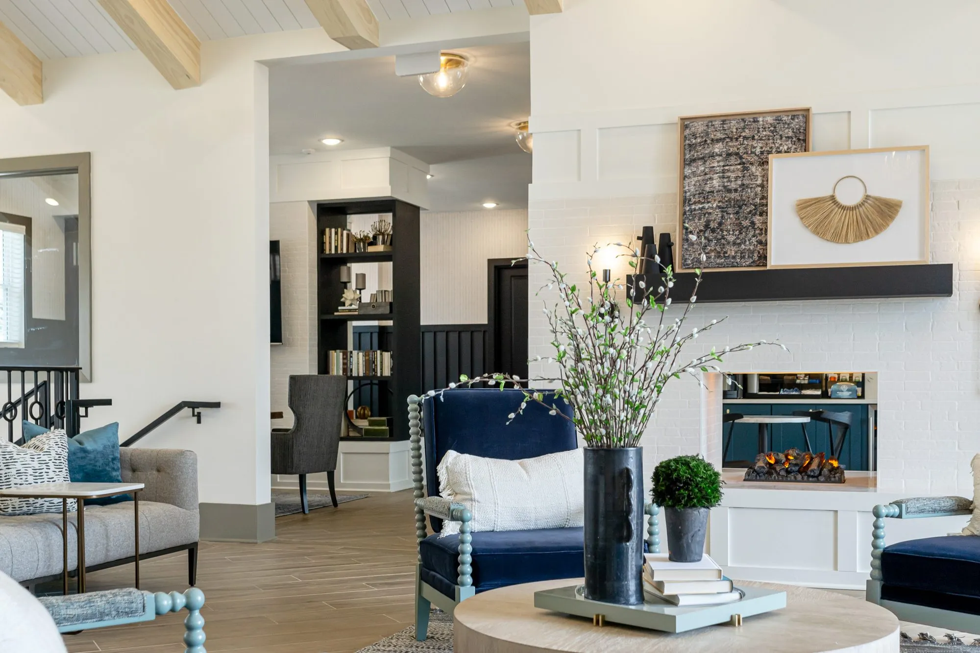 Multifamily amenity lounge with navy spindle chairs and a round wood table before a white brick fireplace, art on the mantel