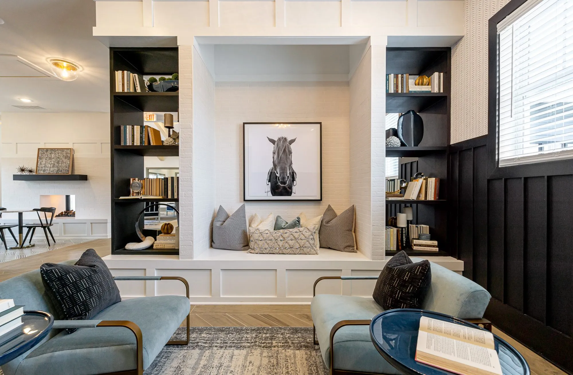 Multifamily amenity library with blue velvet brass-framed chairs facing the bench alcove, a book on a round black side table
