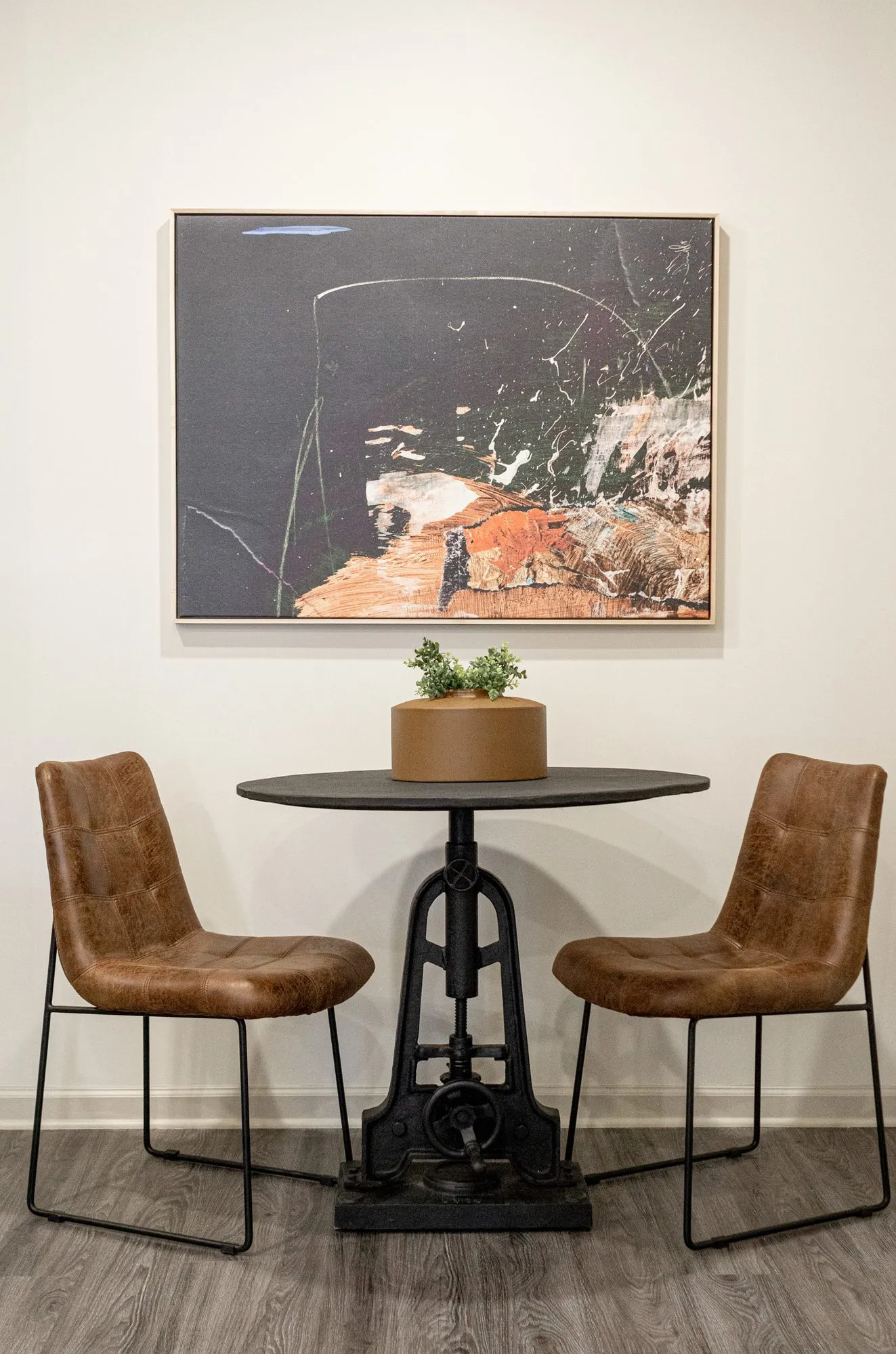 Multifamily interior detail: two tan leather chairs at a black cast iron pedestal table below a framed abstract painting