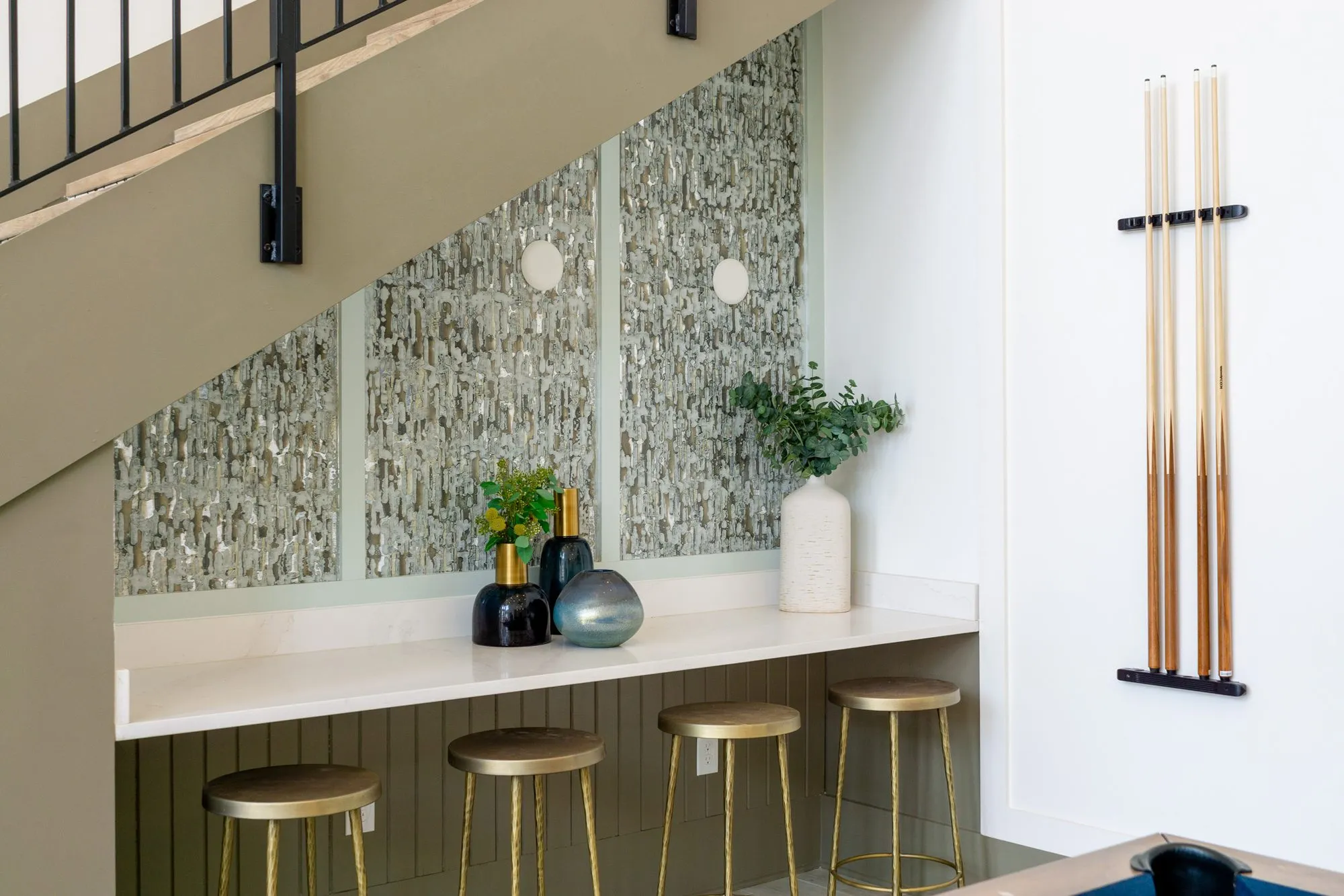 Multifamily amenity counter with brass stools tucked under the stair beside a wall-mounted pool cue rack