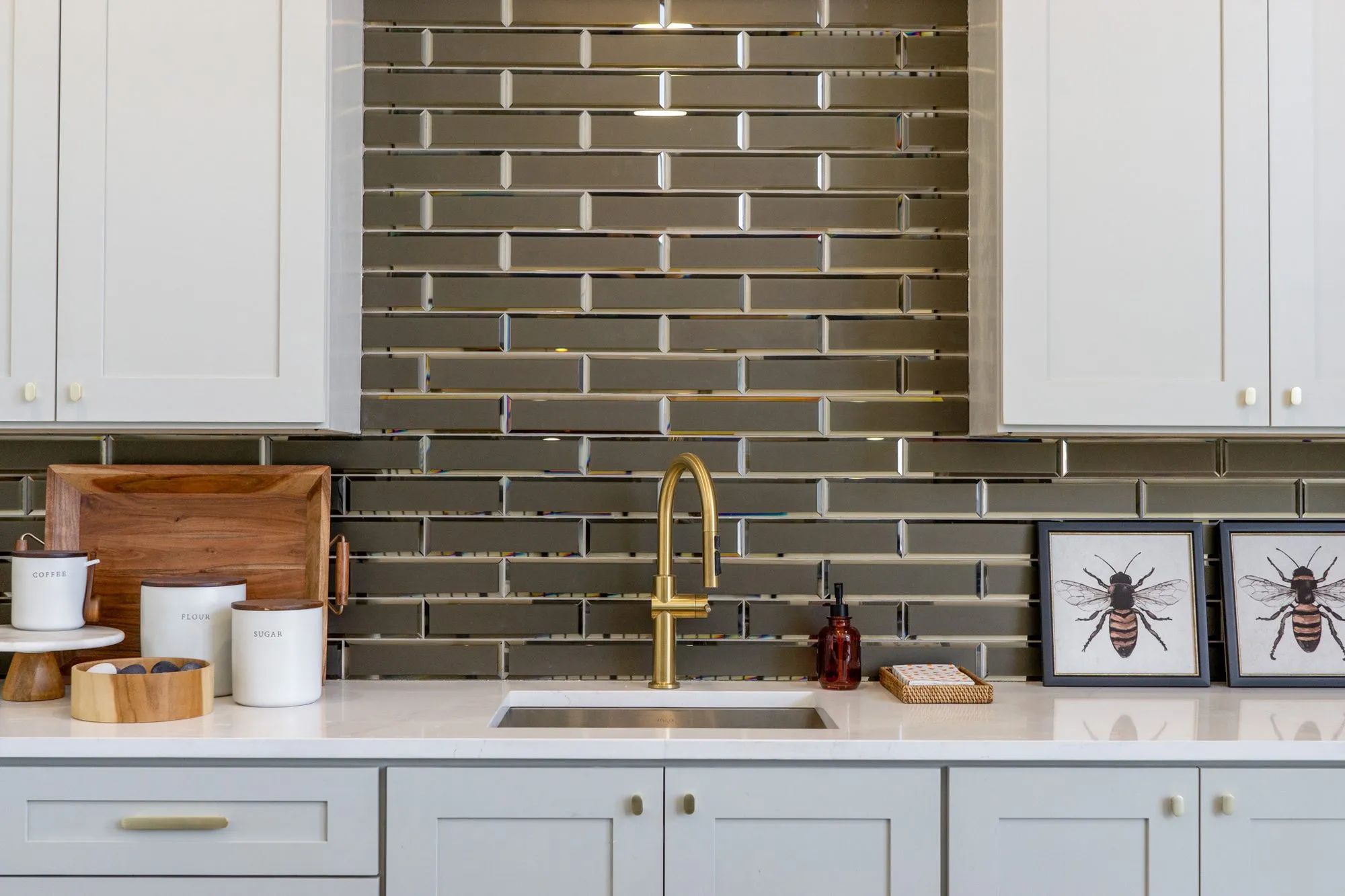 Multifamily interior detail: brass faucet on smoky glass brick tile between grey cabinets, bee prints on the counter