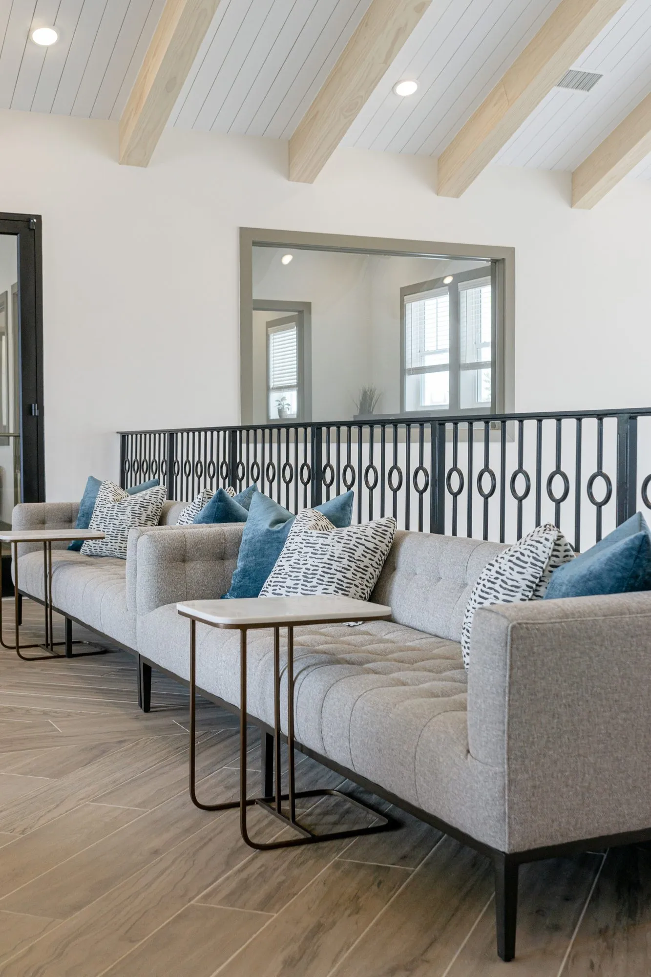 Multifamily amenity mezzanine lounge with two grey tufted sofas, blue pillows and bronze C-shaped tables along the iron rail