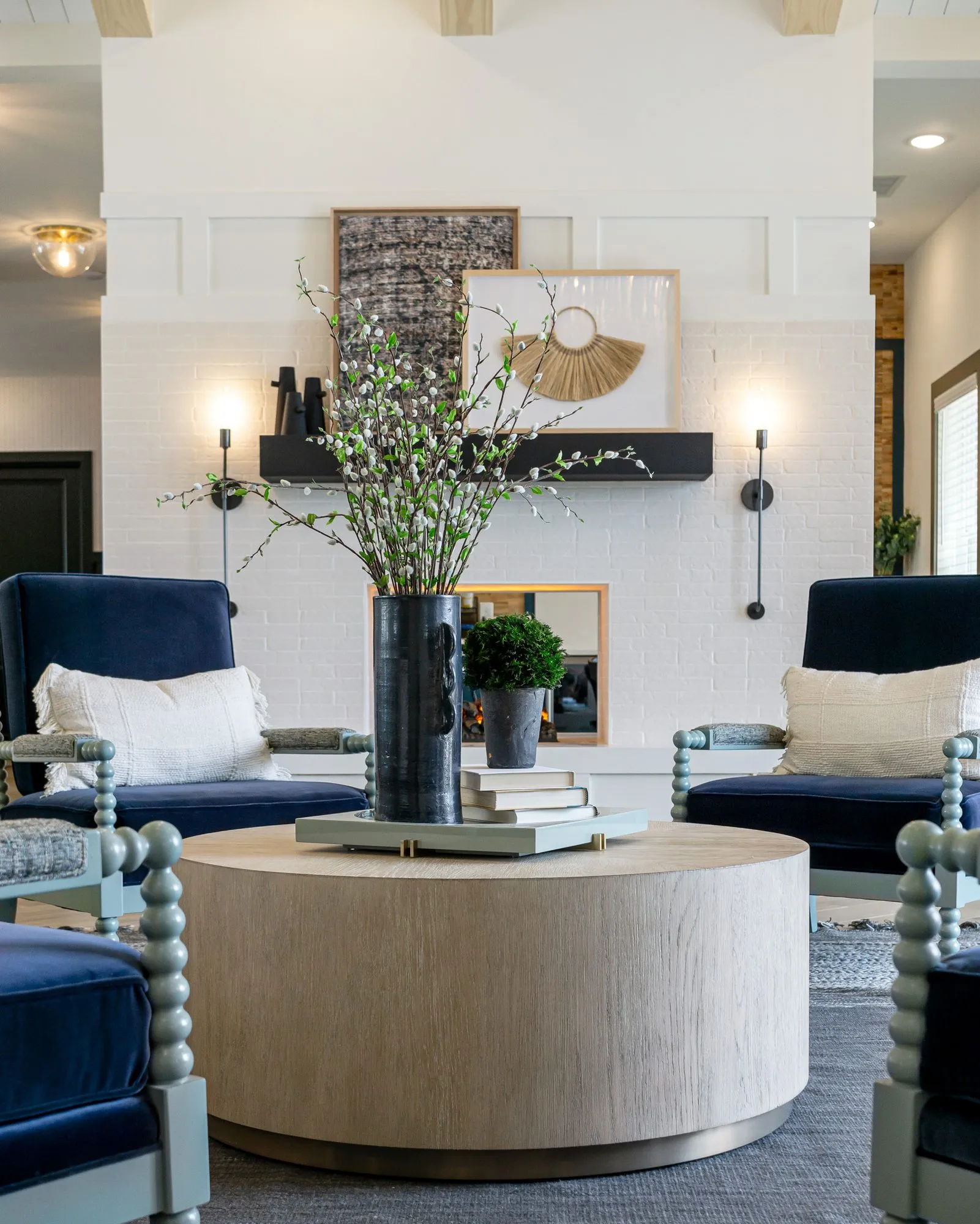 Multifamily interior detail: wood drum table with flowering branches between spindle chairs, white brick fireplace behind