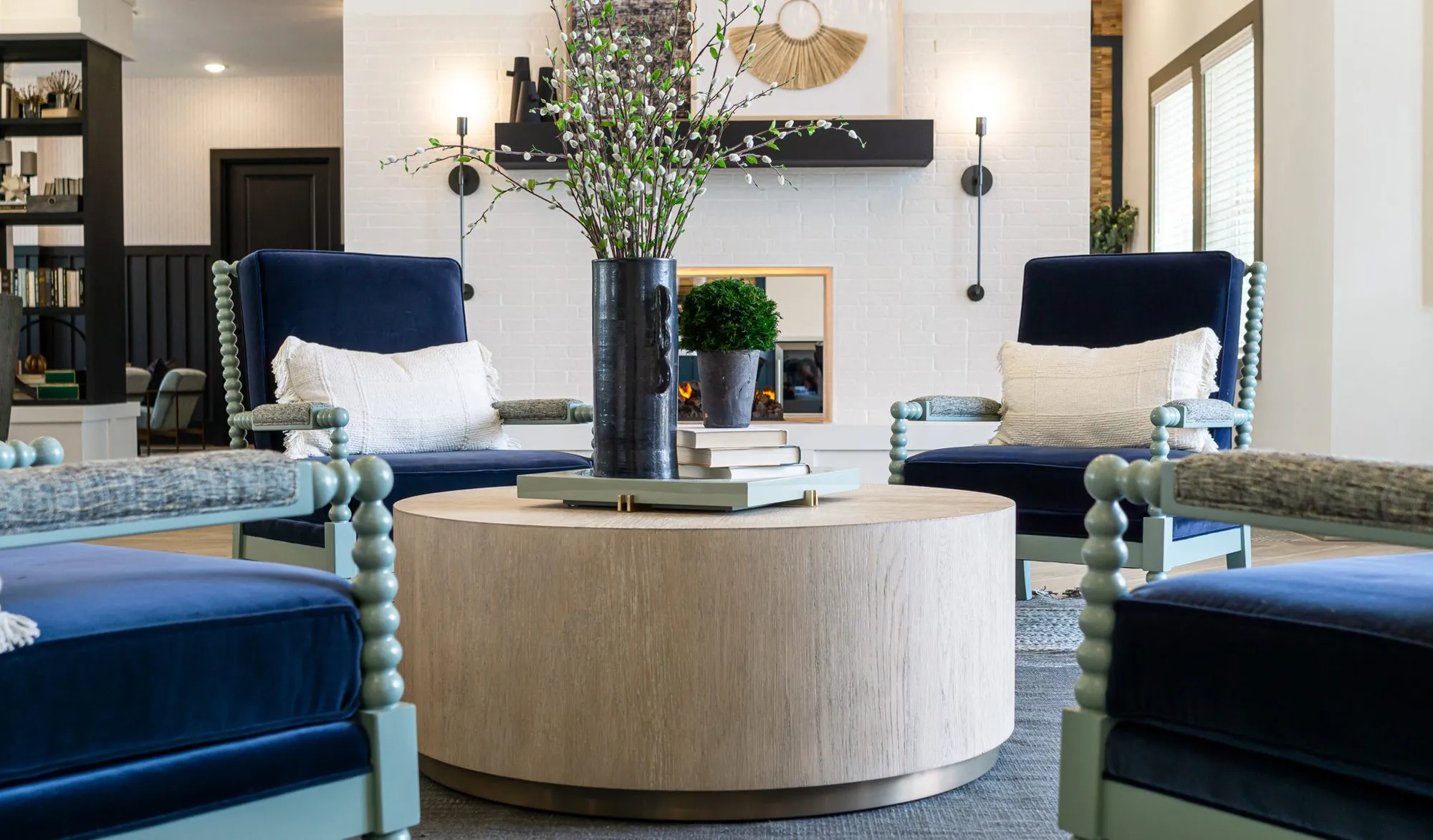 Multifamily amenity lounge, low view across navy velvet spindle chairs to a wood drum table and white brick fireplace