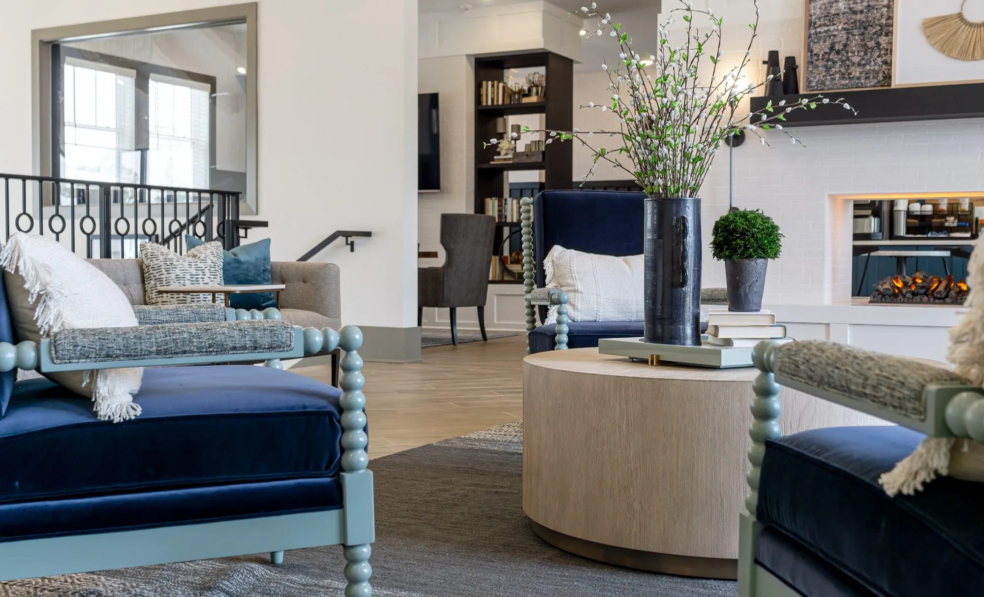 Multifamily amenity lounge with navy spindle chairs at a drum table, the fireplace, stair rail and bookcases beyond