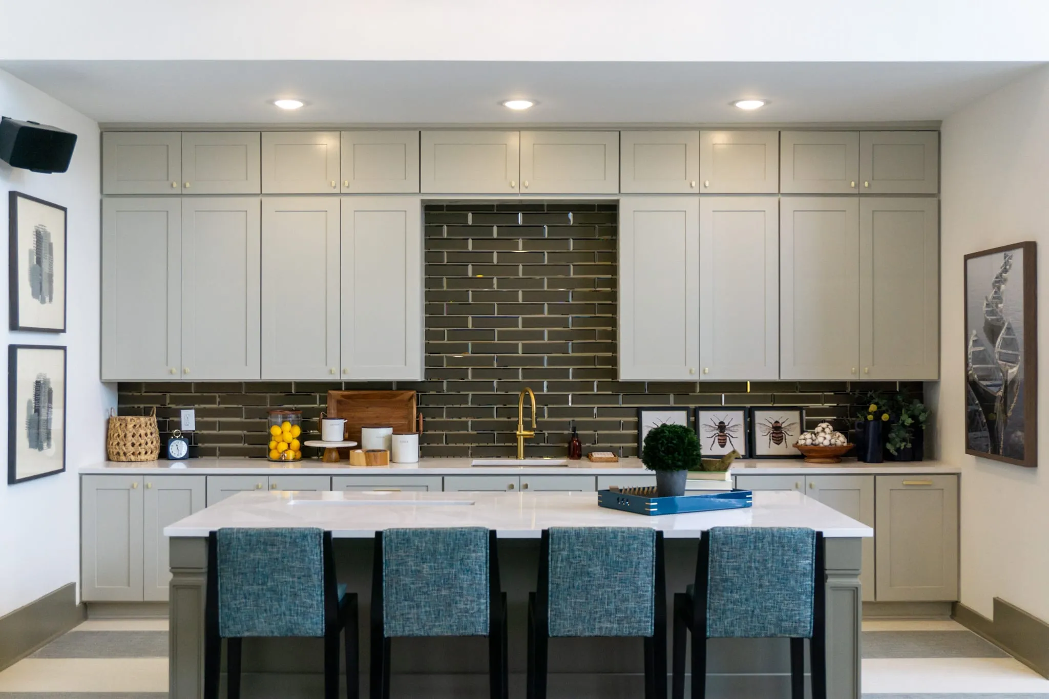 Multifamily amenity clubhouse kitchen with olive subway tile behind pale cabinetry and teal upholstered stools at the island