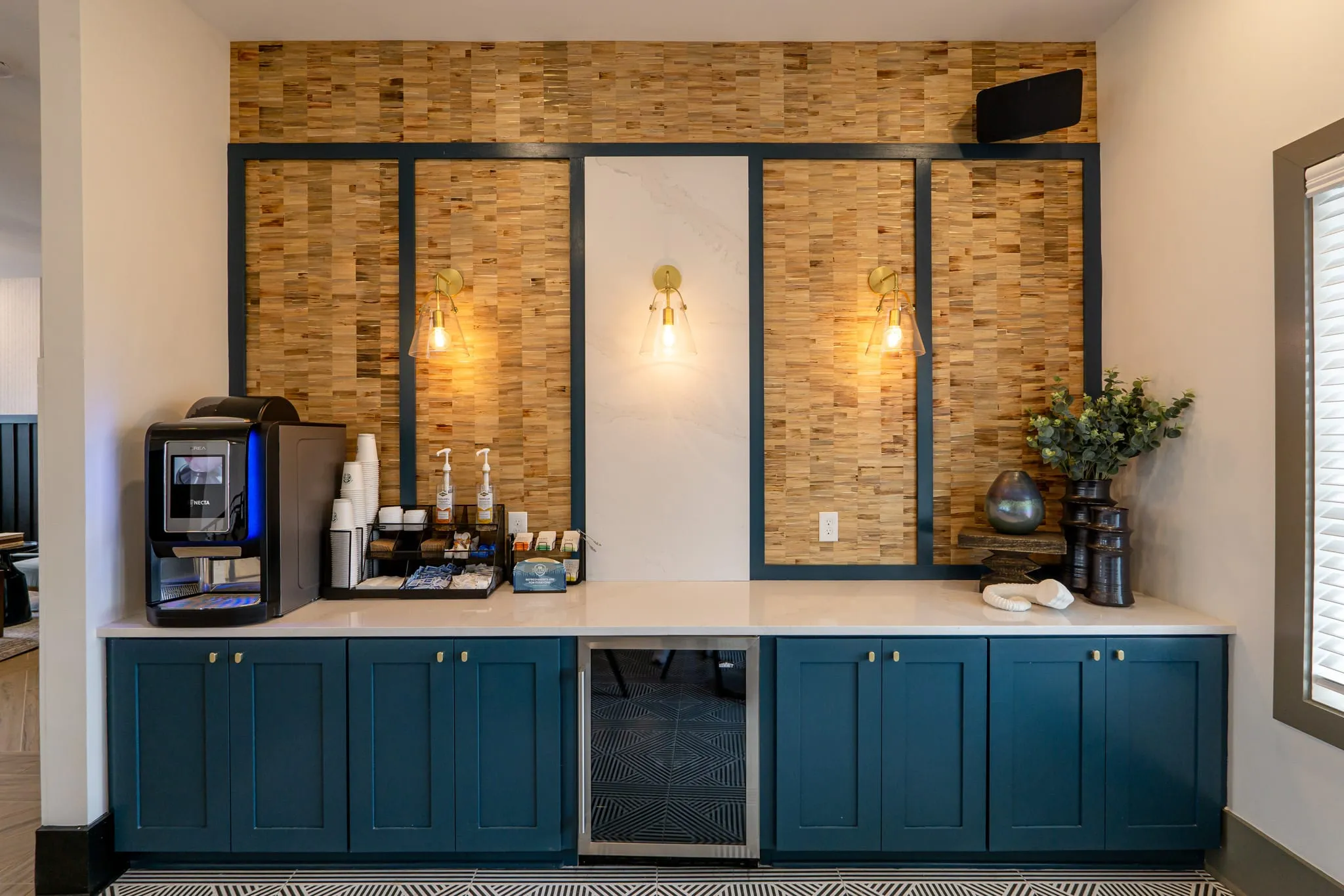 Multifamily amenity coffee bar with teal cabinetry below a wood brick backsplash framed in black steel with exposed bulbs