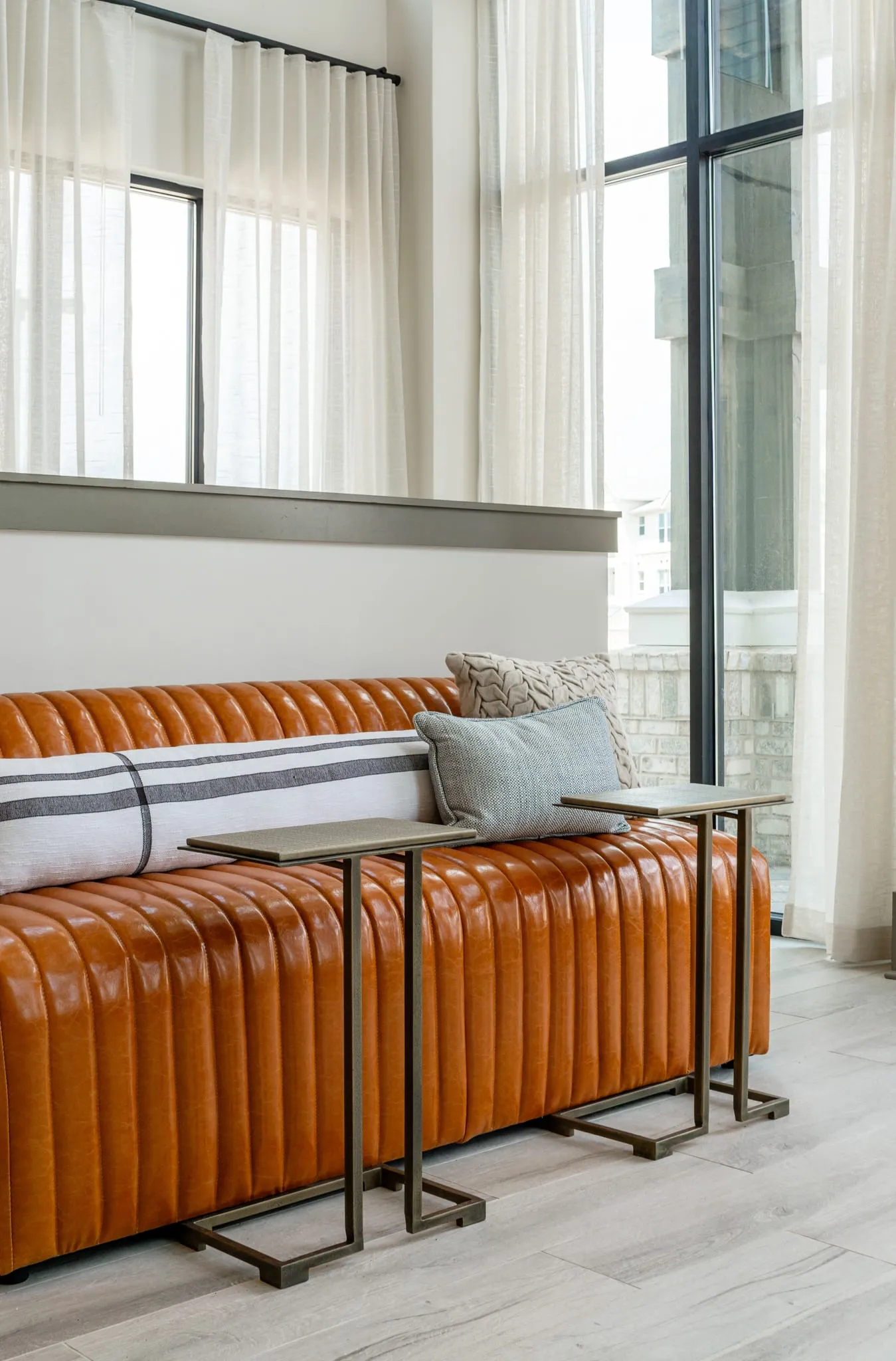 Multifamily amenity lounge with a cognac channel tufted leather bench and striped pillow below tall curtained windows