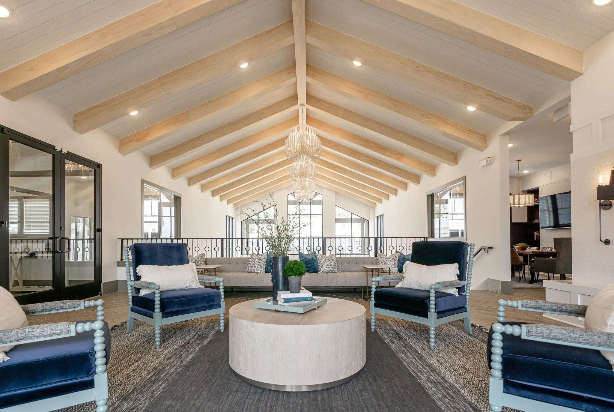 Multifamily amenity clubhouse lounge with navy spindle settees around an upholstered round table under a vaulted beam ceiling