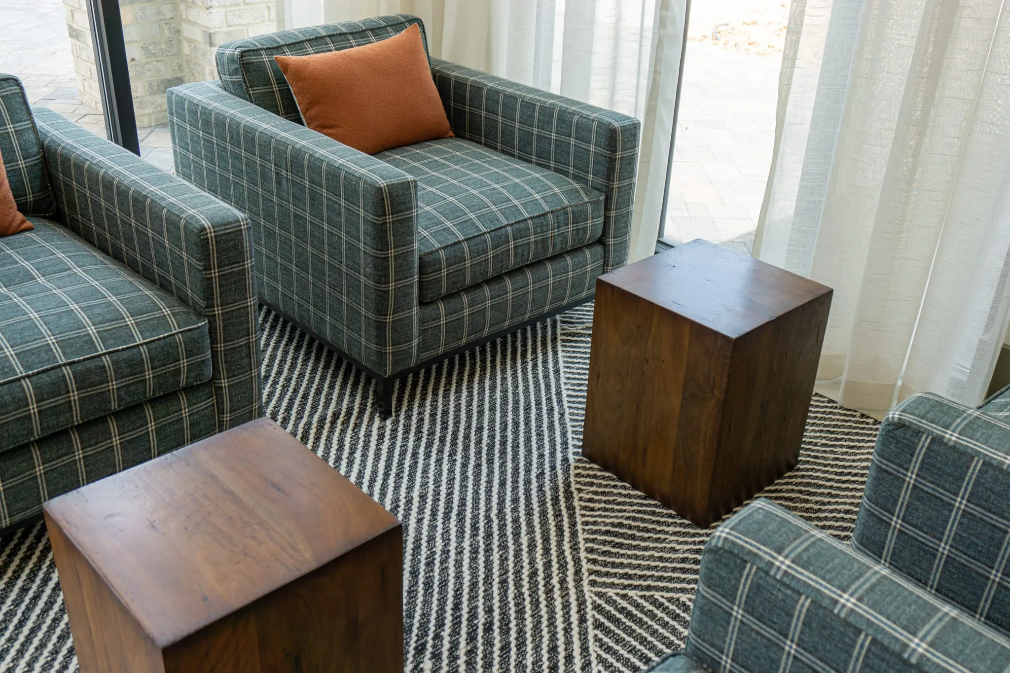 Multifamily amenity lounge with green plaid armchairs and walnut cube tables on a striped rug beside sheer curtains