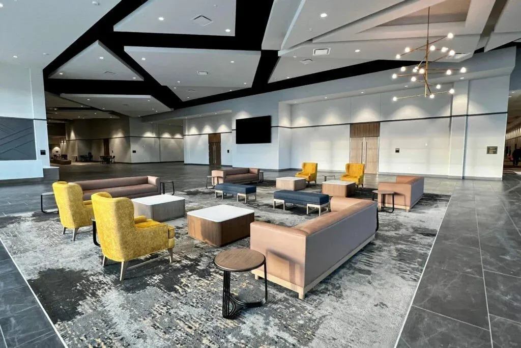Hotel prefunction lounge with yellow armchairs and blush sofas on a patterned carpet, under a black geometric ceiling