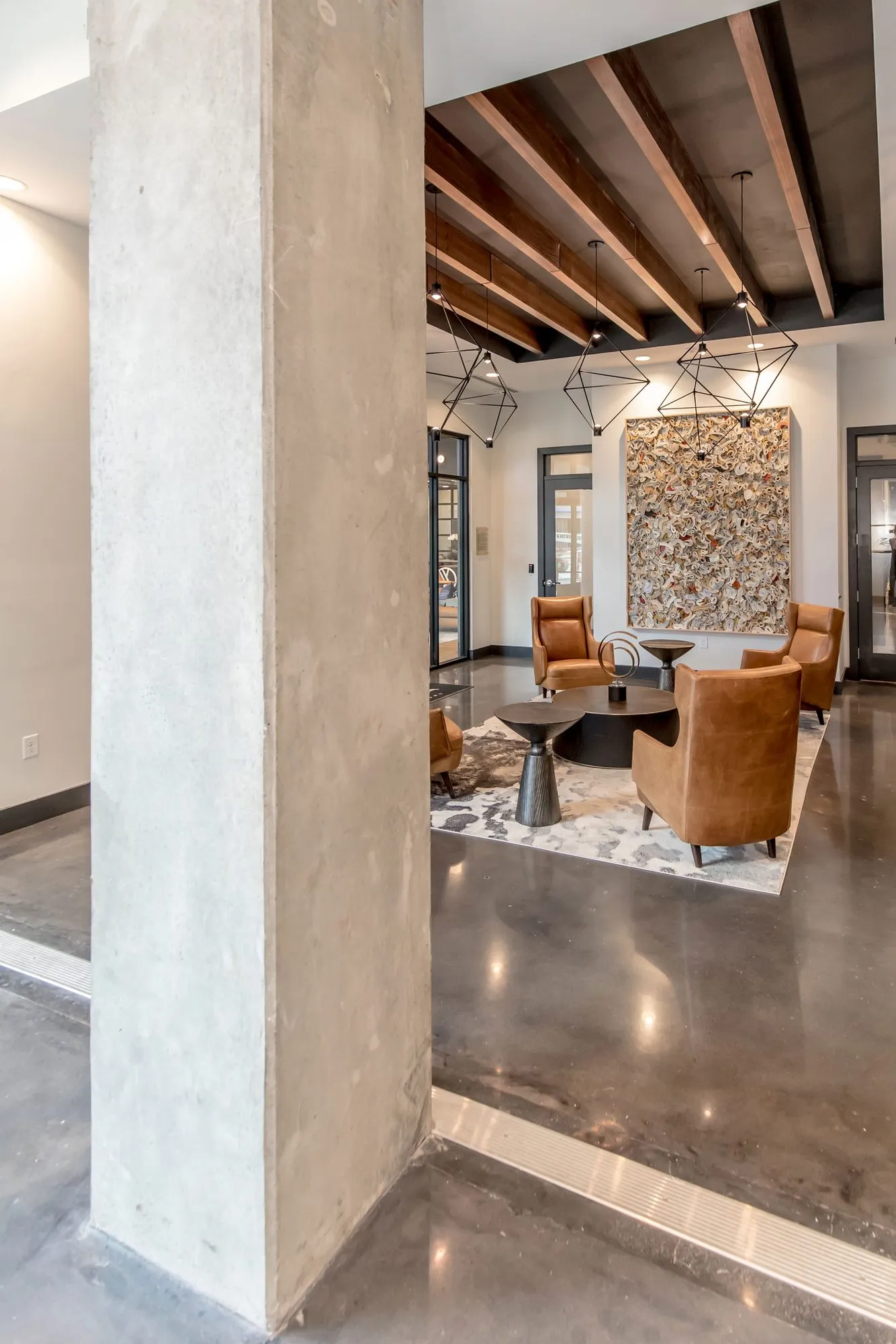 Multifamily amenity lounge with tan leather armchairs under wood beams and wire geometric pendants beyond a concrete column