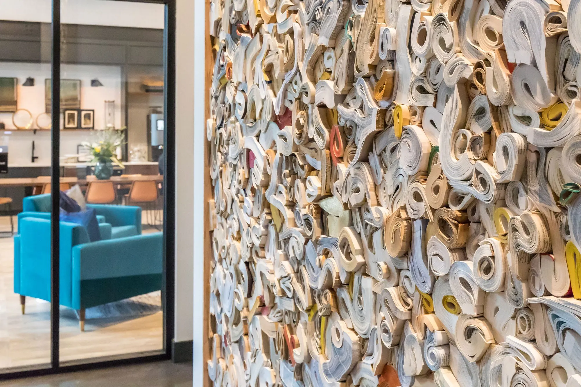 Multifamily amenity lounge with a teal sofa seen through a glass door beside a sculptural wall of rolled paper