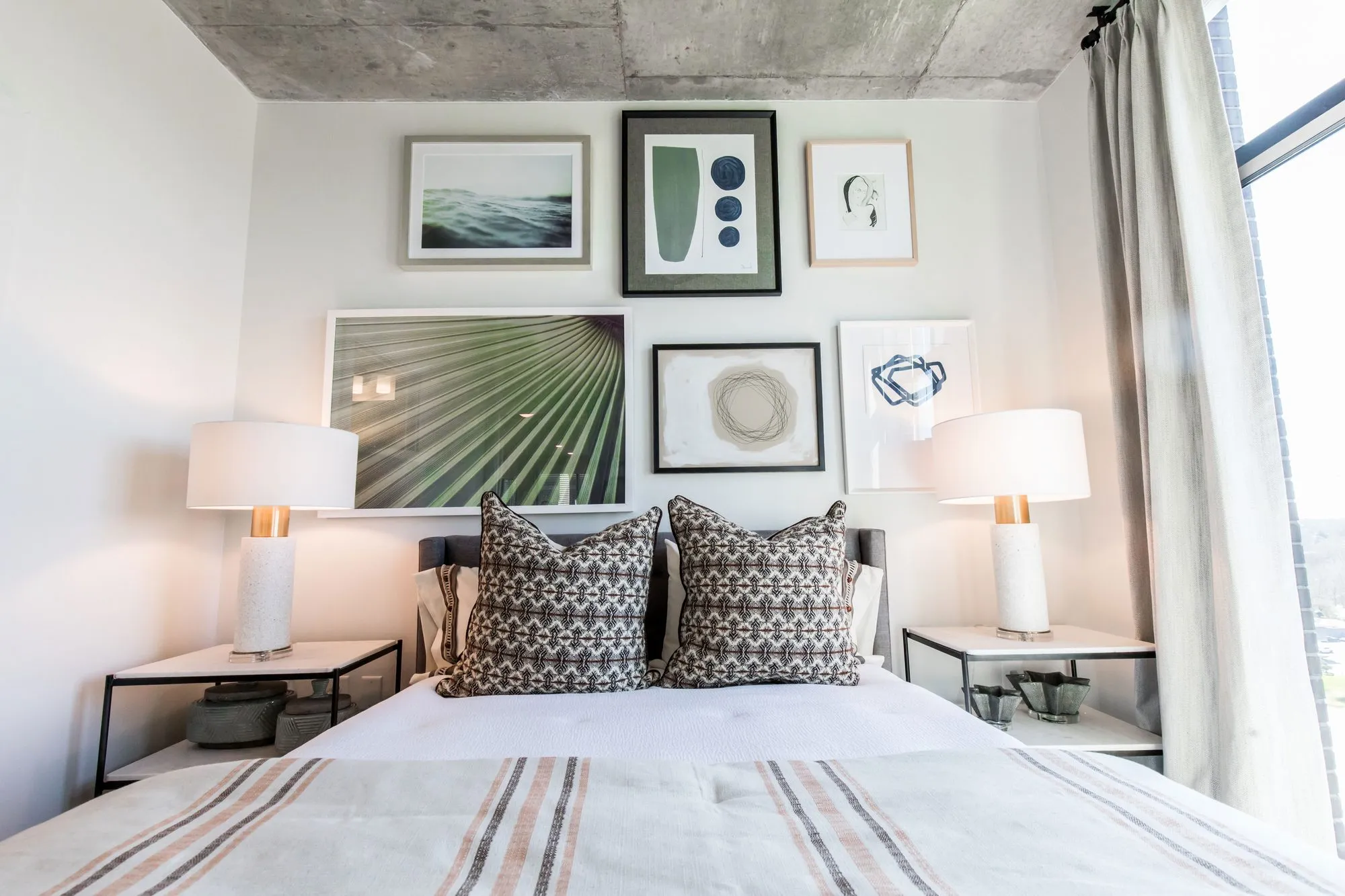Multifamily model apartment bedroom with striped bedding, two white table lamps and a gallery wall under a concrete ceiling