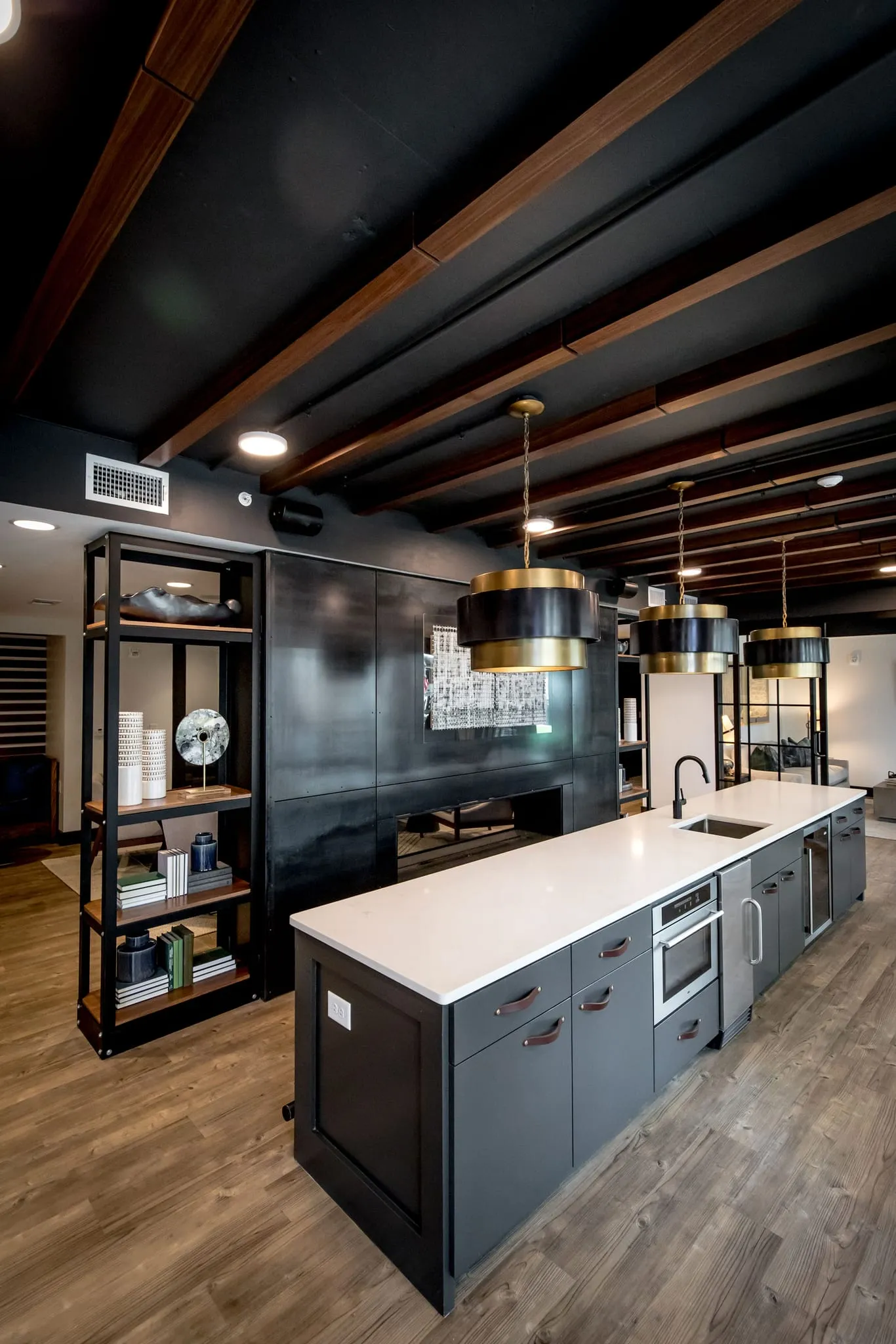 Multifamily amenity clubhouse kitchen with dark cabinetry and white counters under black beams with brass pendants