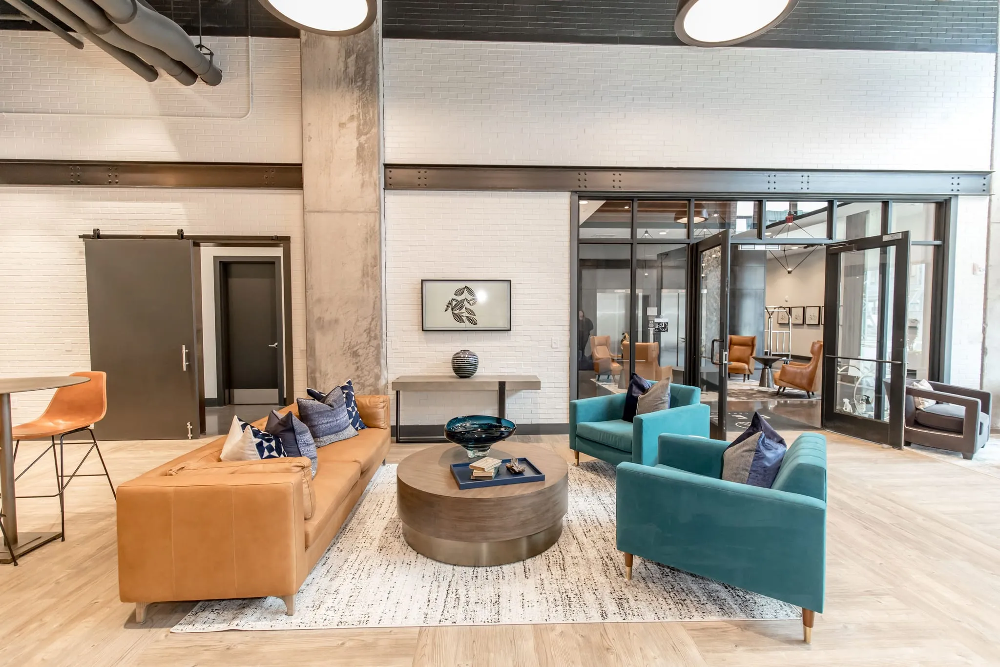 Multifamily amenity clubroom with a camel leather sofa facing teal armchairs around a wood drum table by a sliding barn door