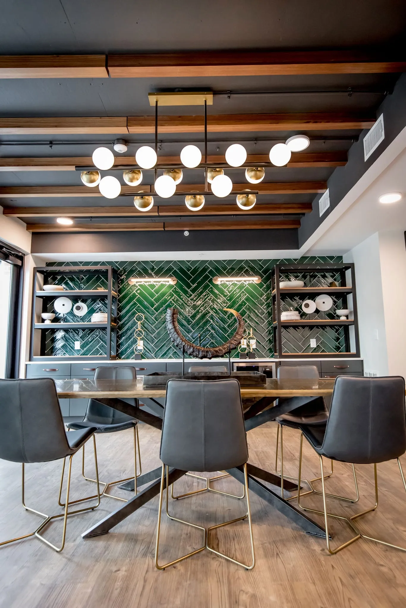 Multifamily amenity private dining room with grey chairs below a brass globe chandelier, the green tile bar behind